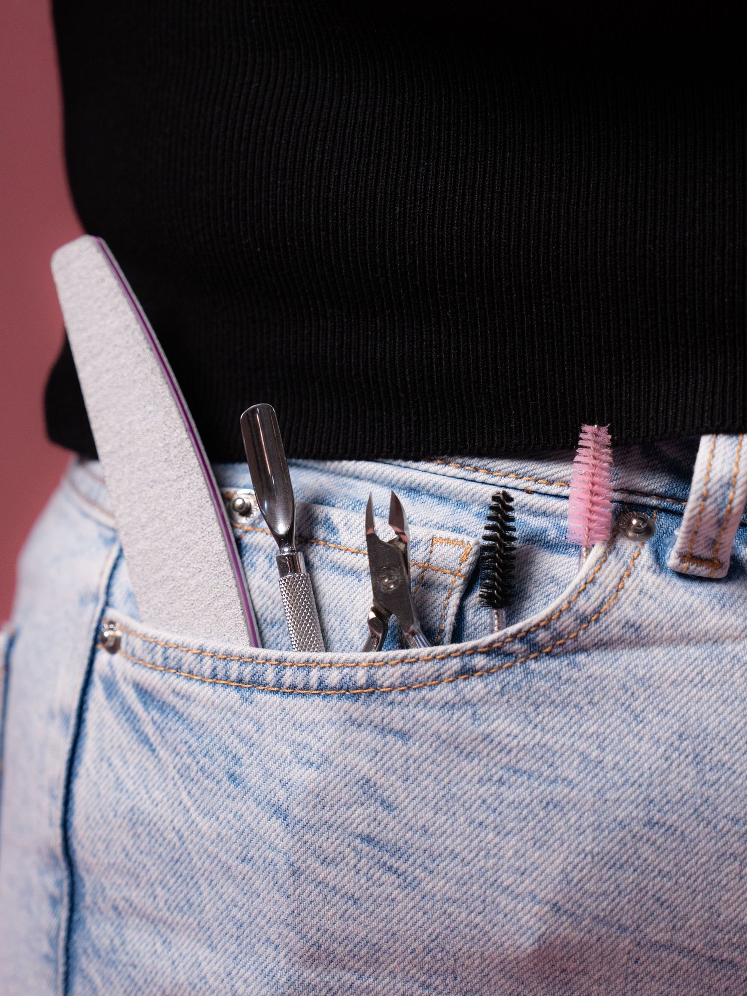 Various makeup brushes and tools peek out from a light wash denim jeans pocket against a pink background.