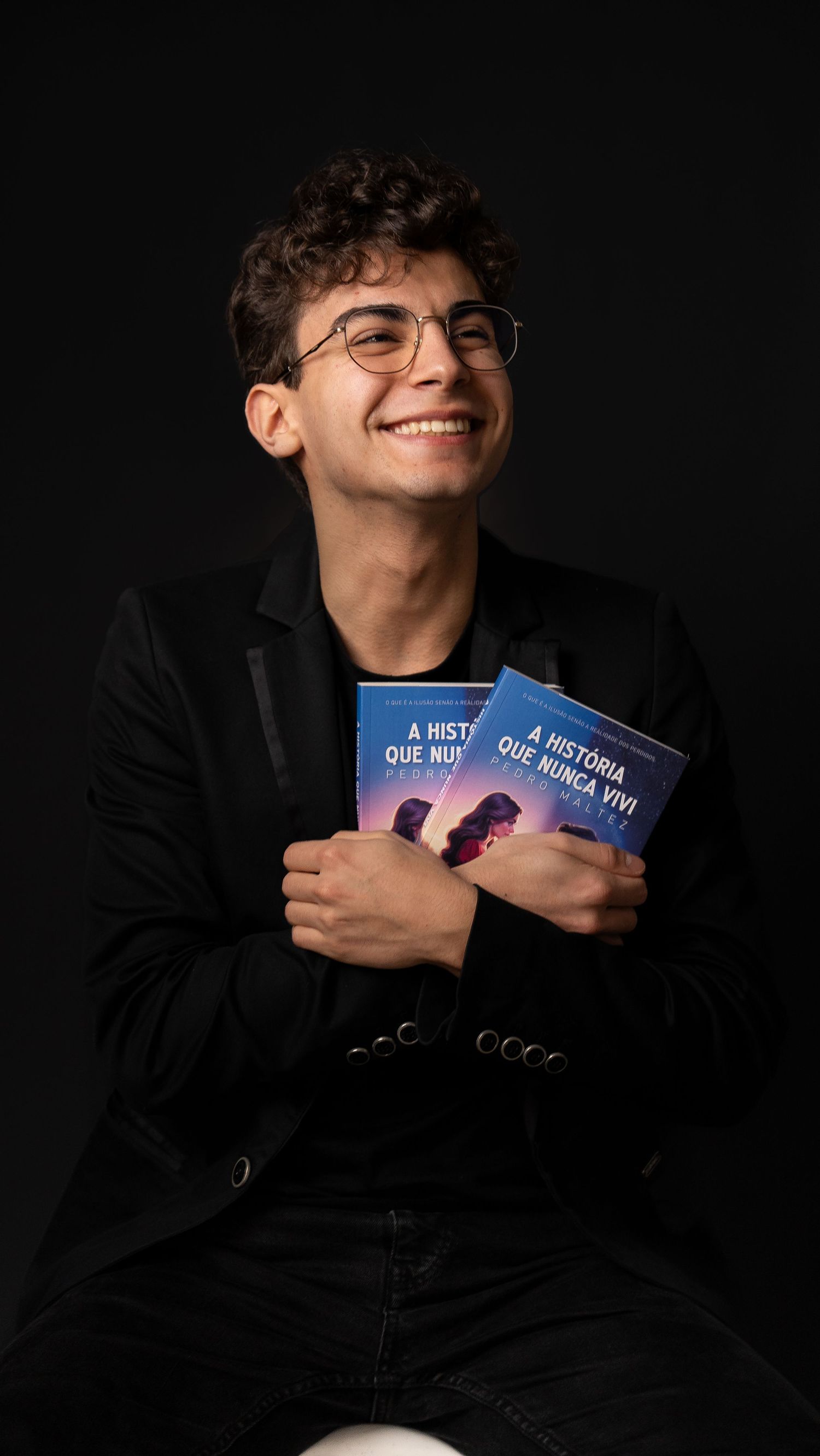 Person in black attire holds multiple books while smiling against dark backdrop.