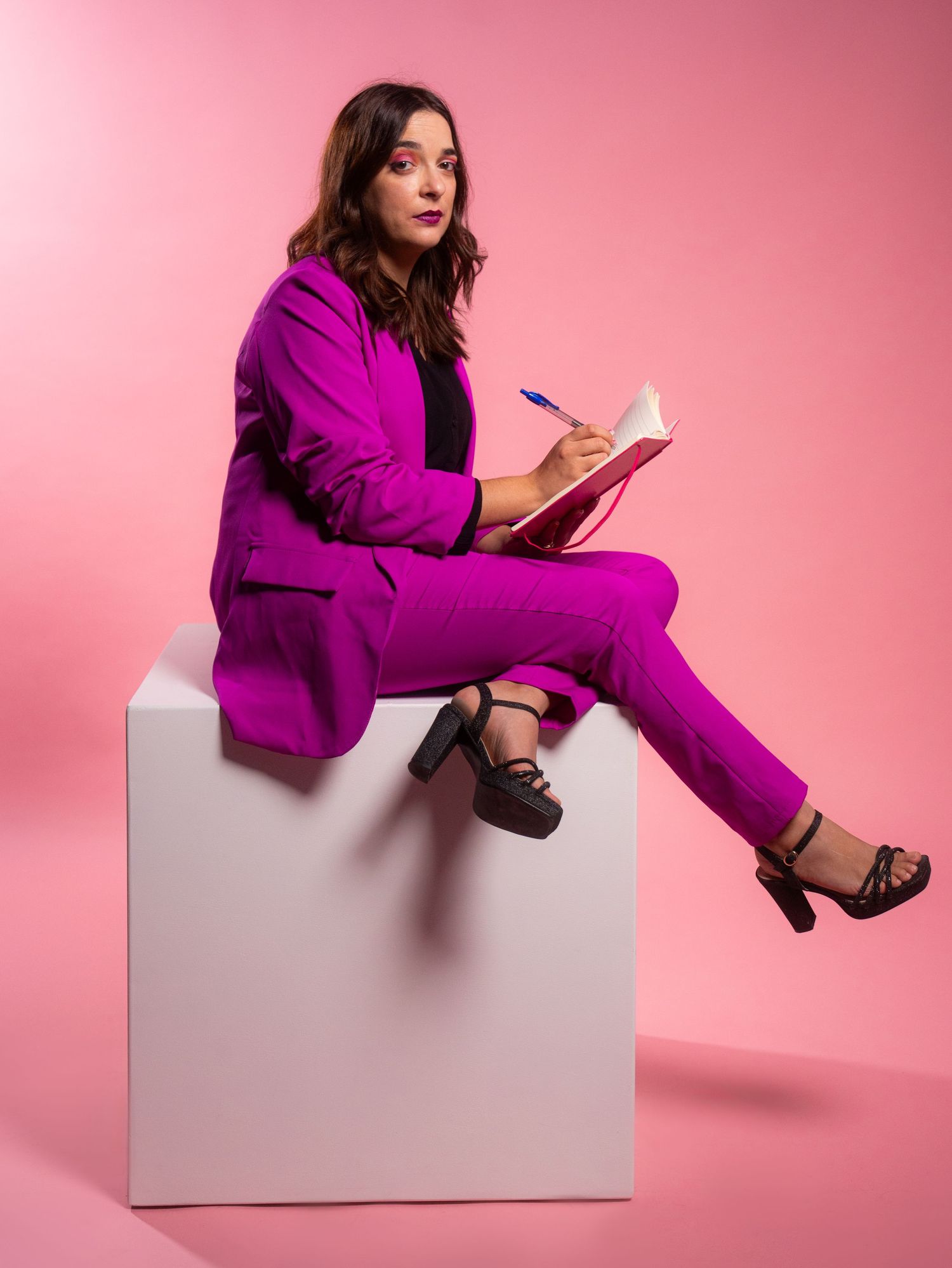 Stylish figure in purple suit poses on white cube against pink studio background.