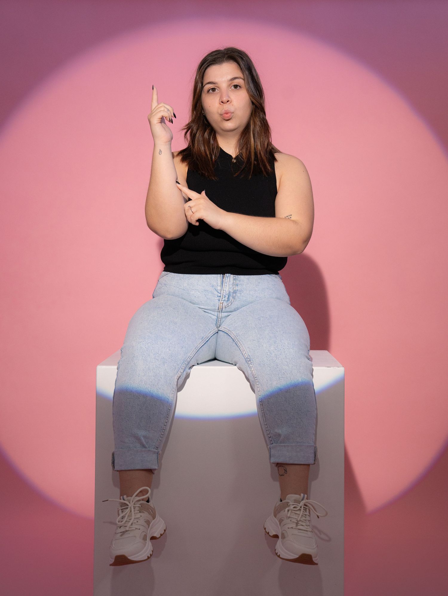 A person in black top and jeans sits on white platform with spotlight effect.