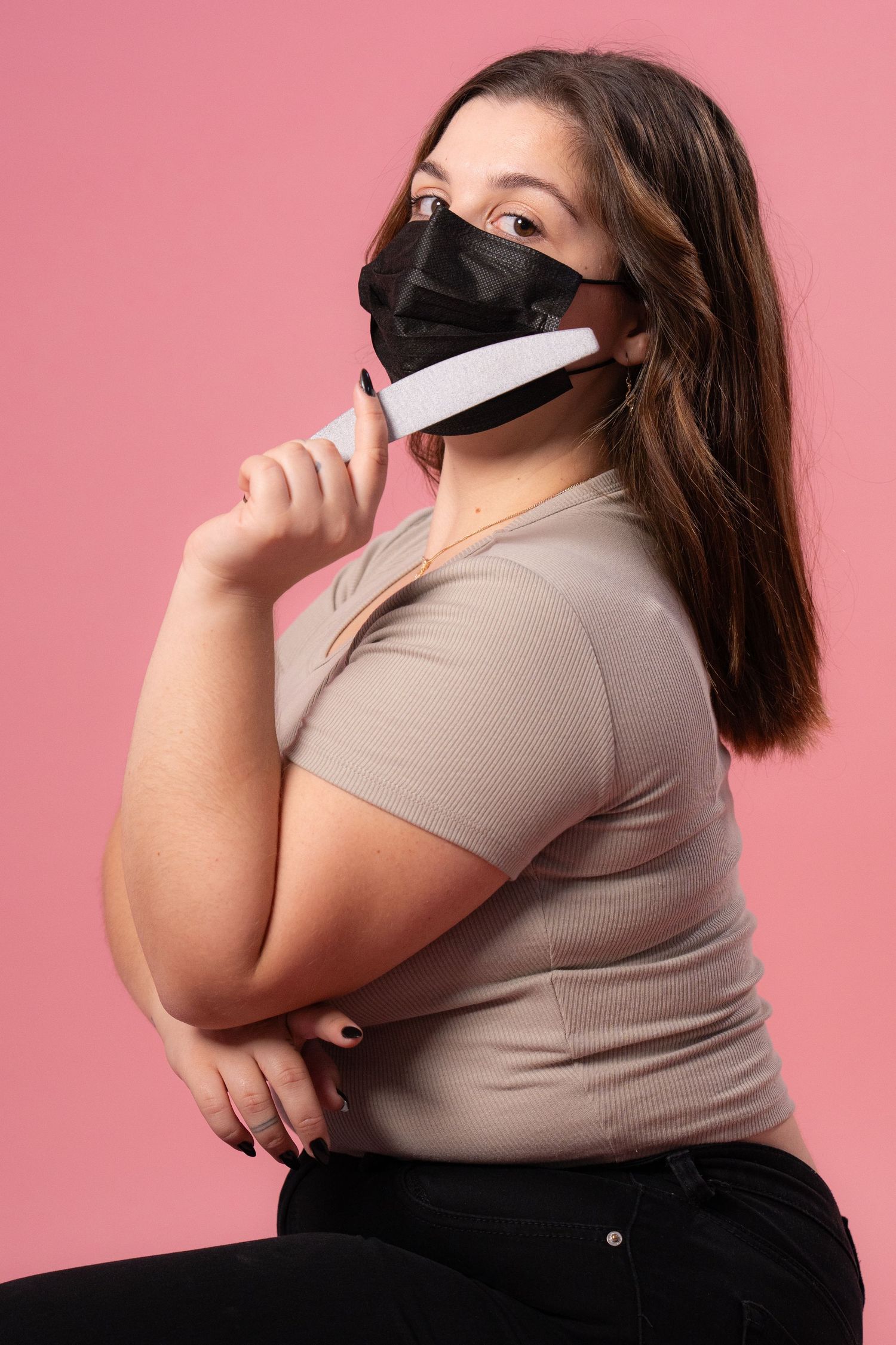 Model wearing black face mask poses with nail file against pink background.