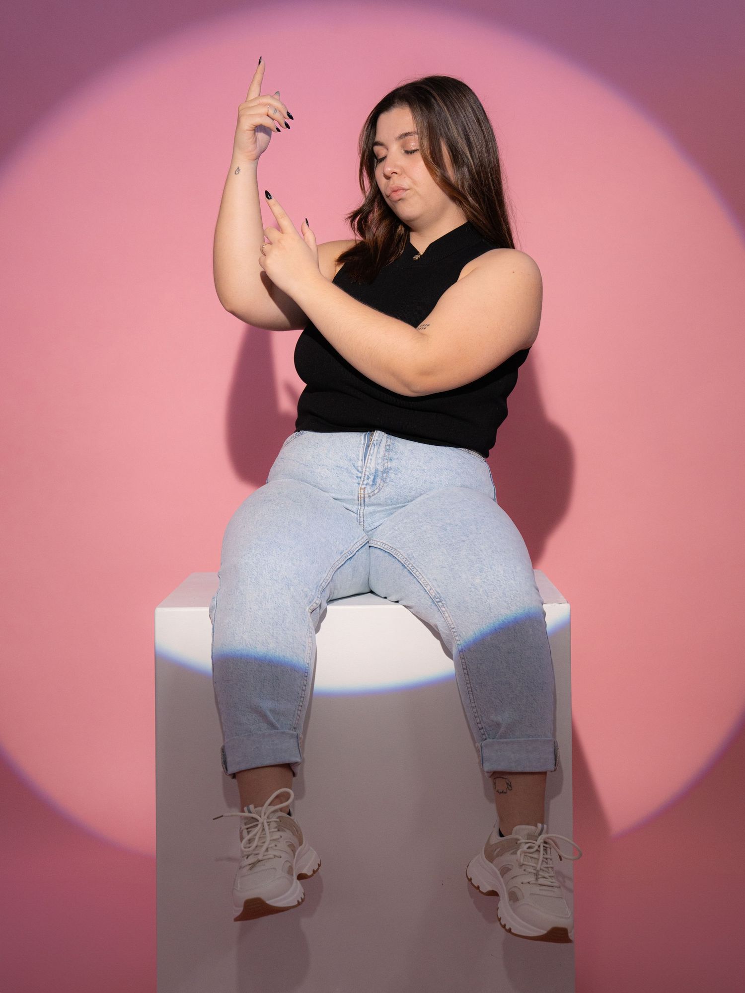 A person poses playfully while sitting on white platform against pink background.