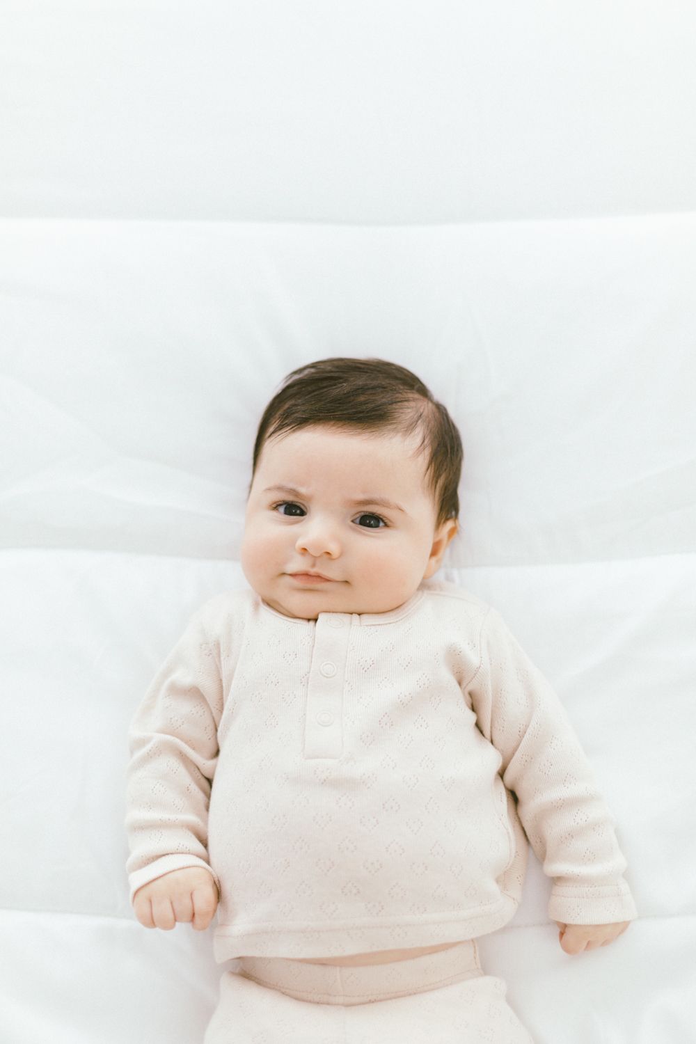 Baby in cream colored outfit smiling while lying on white bedding.