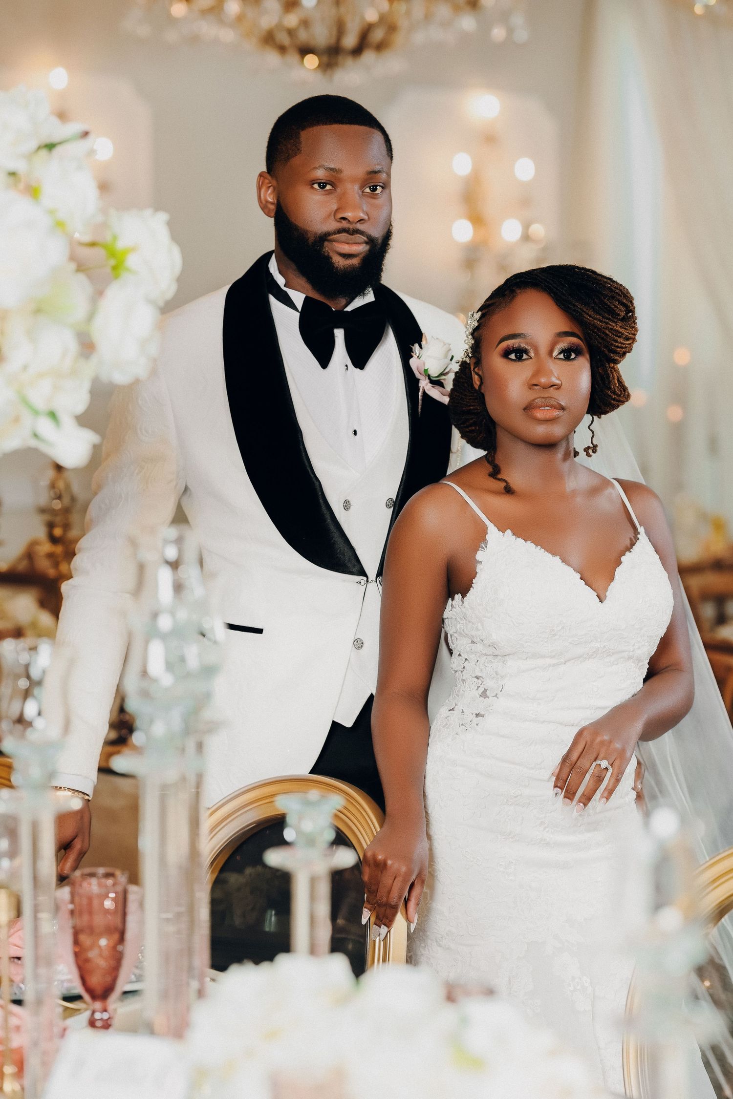 Elegant bride and groom pose together at decorated reception table with white floral arrangements and candles.