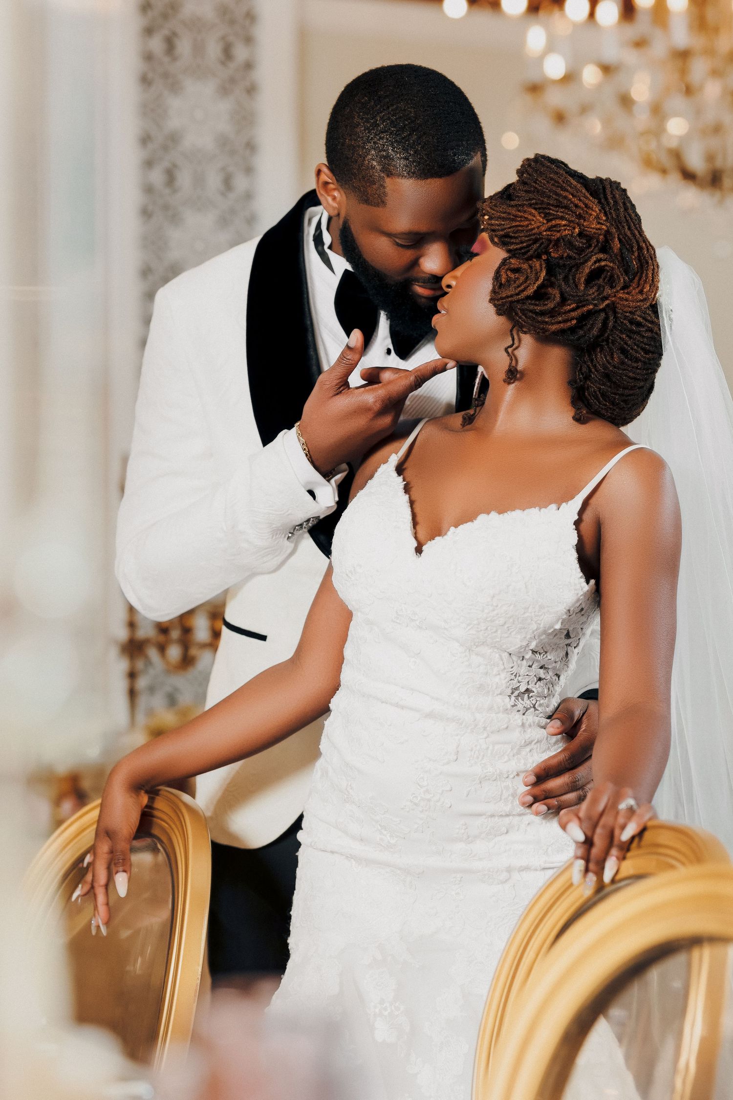 Groom in white tuxedo leans in to kiss bride in lace gown beside ornate gold chair with chandelier backdrop.