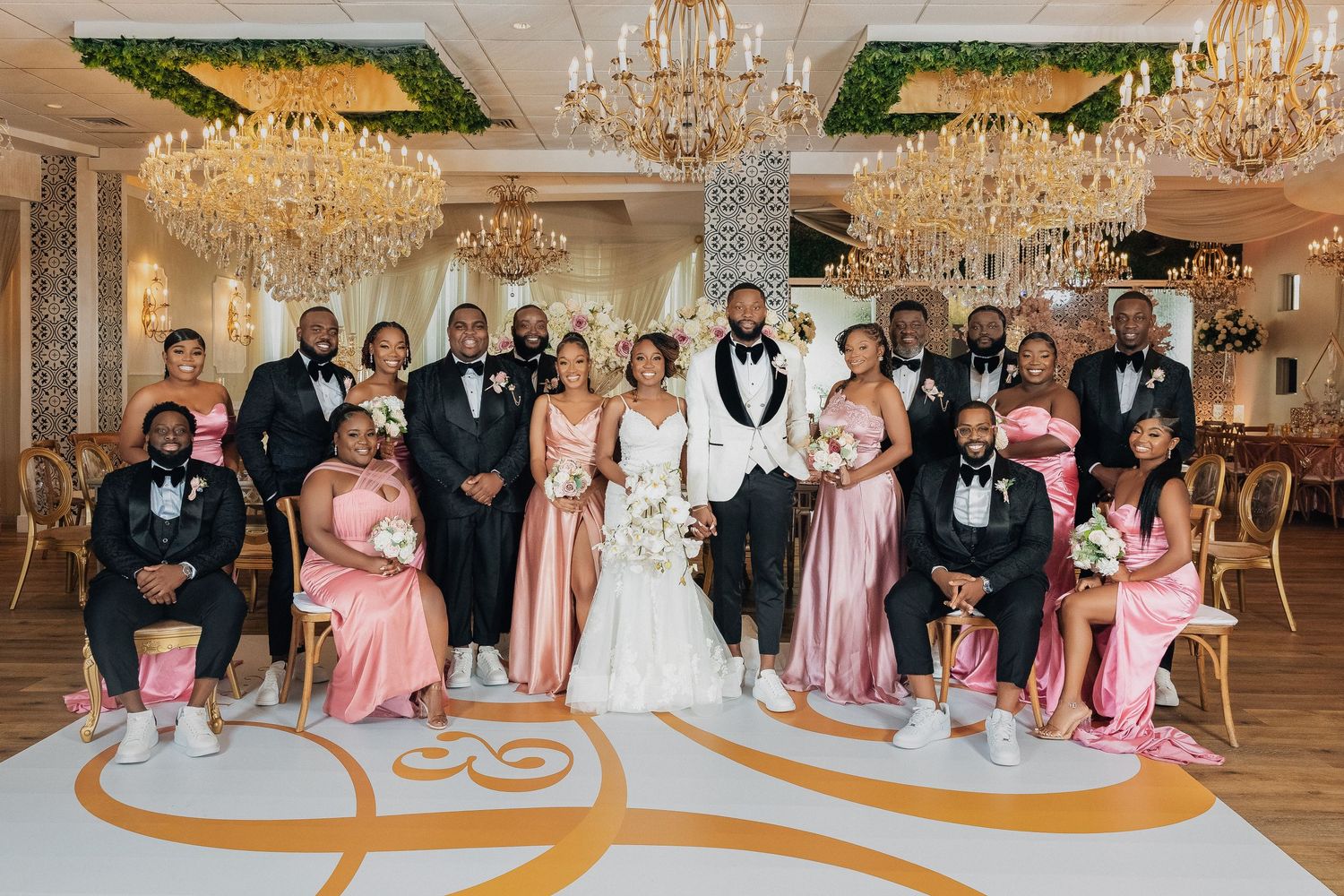 Wedding party poses in pink bridesmaid dresses and black tuxedos under grand chandeliers in elegant ballroom.