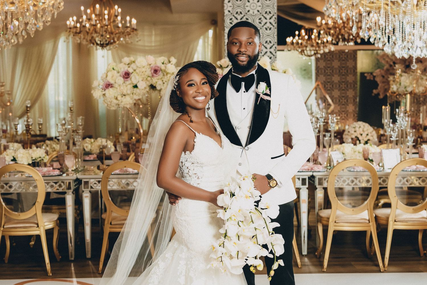 Bride and groom smile together in elegant gold reception venue with white floral centerpieces and chandeliers.