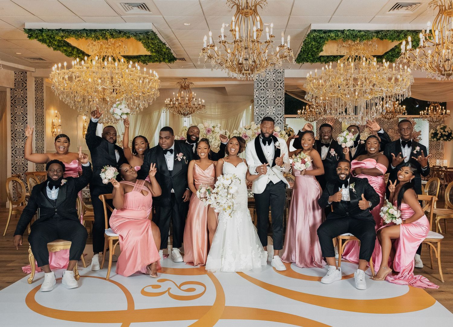 Joyful wedding party celebrates on dance floor under chandeliers with pink bridesmaids and black groomsmen attire.