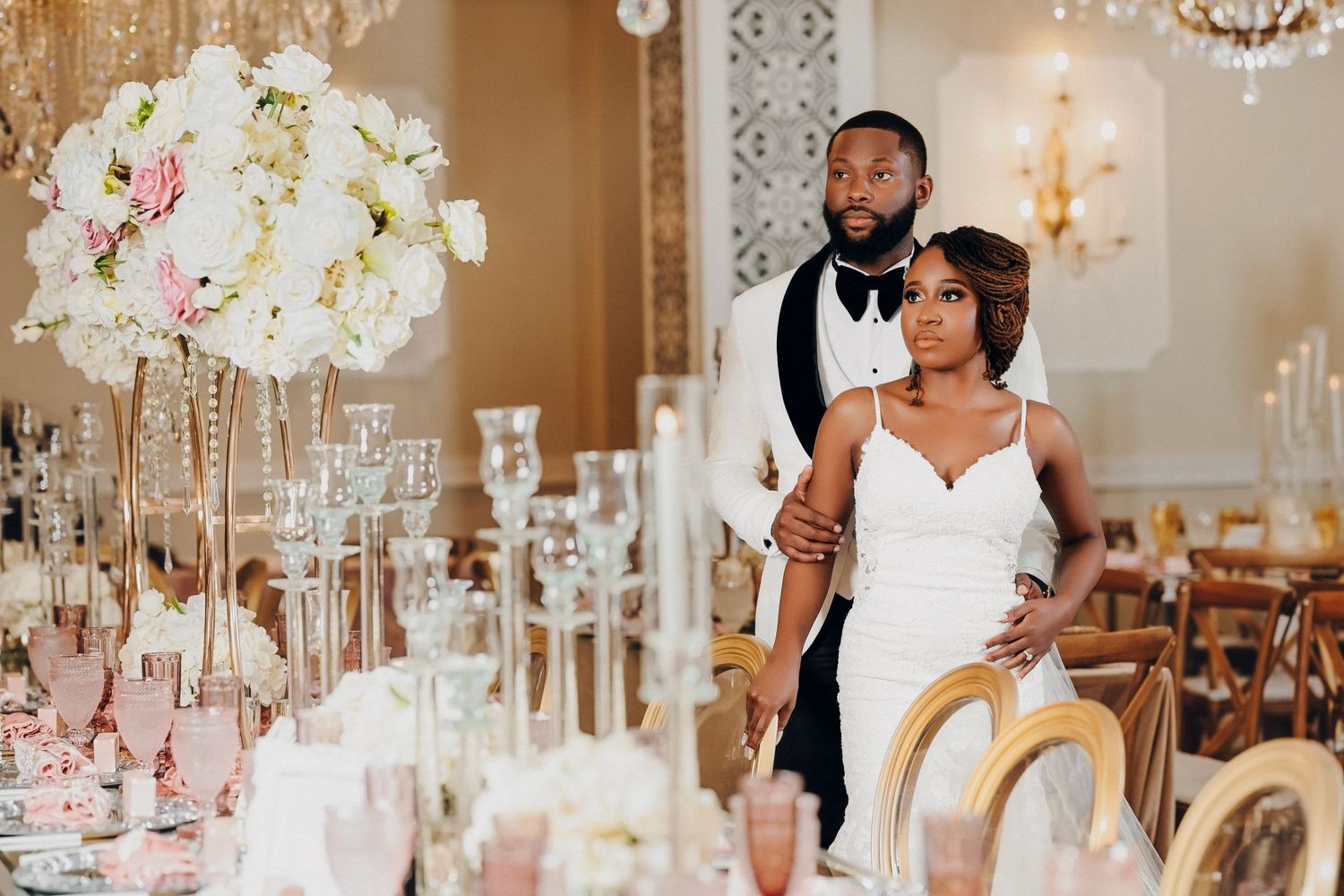 Elegant wedding couple poses in luxurious gold reception hall with tall floral centerpieces and crystal candelabras.