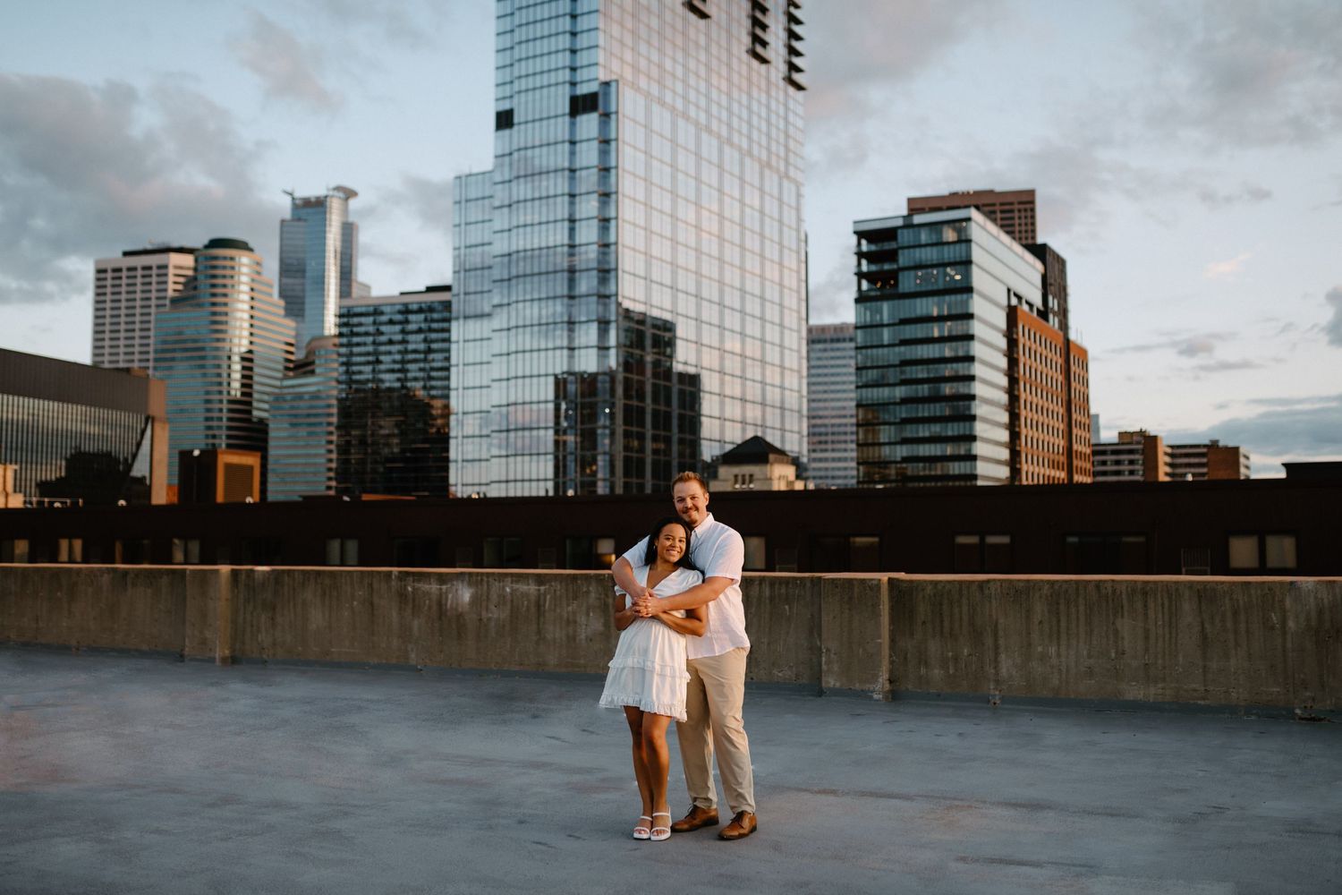 A couple dances on an urban rooftop with modern glass buildings in the background at sunset.