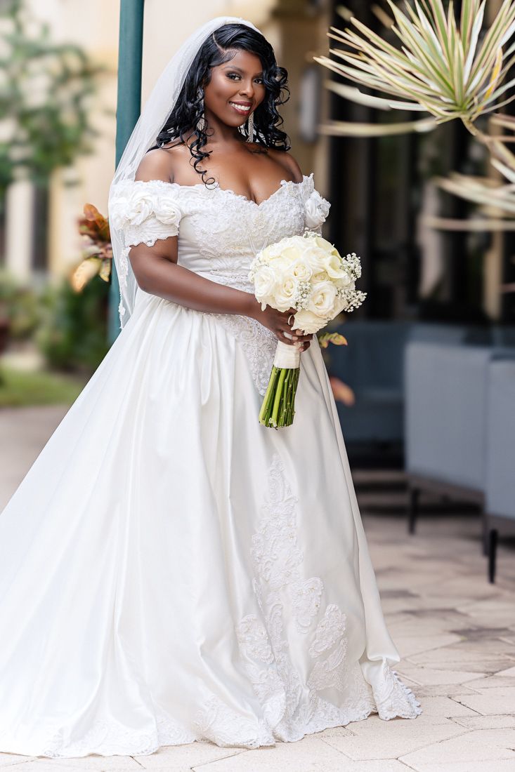 Elegant bride in off-shoulder white gown holds classic white bouquet on outdoor patio.