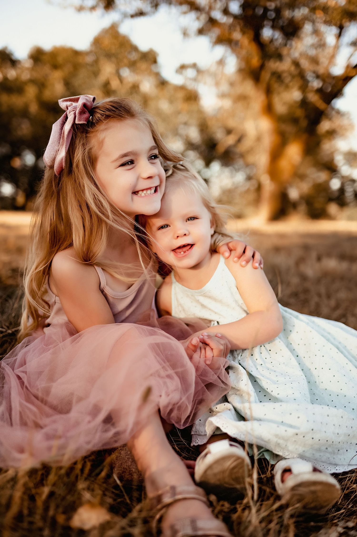 Two children in pink and white dresses share a sweet moment together in a golden sunlit field.