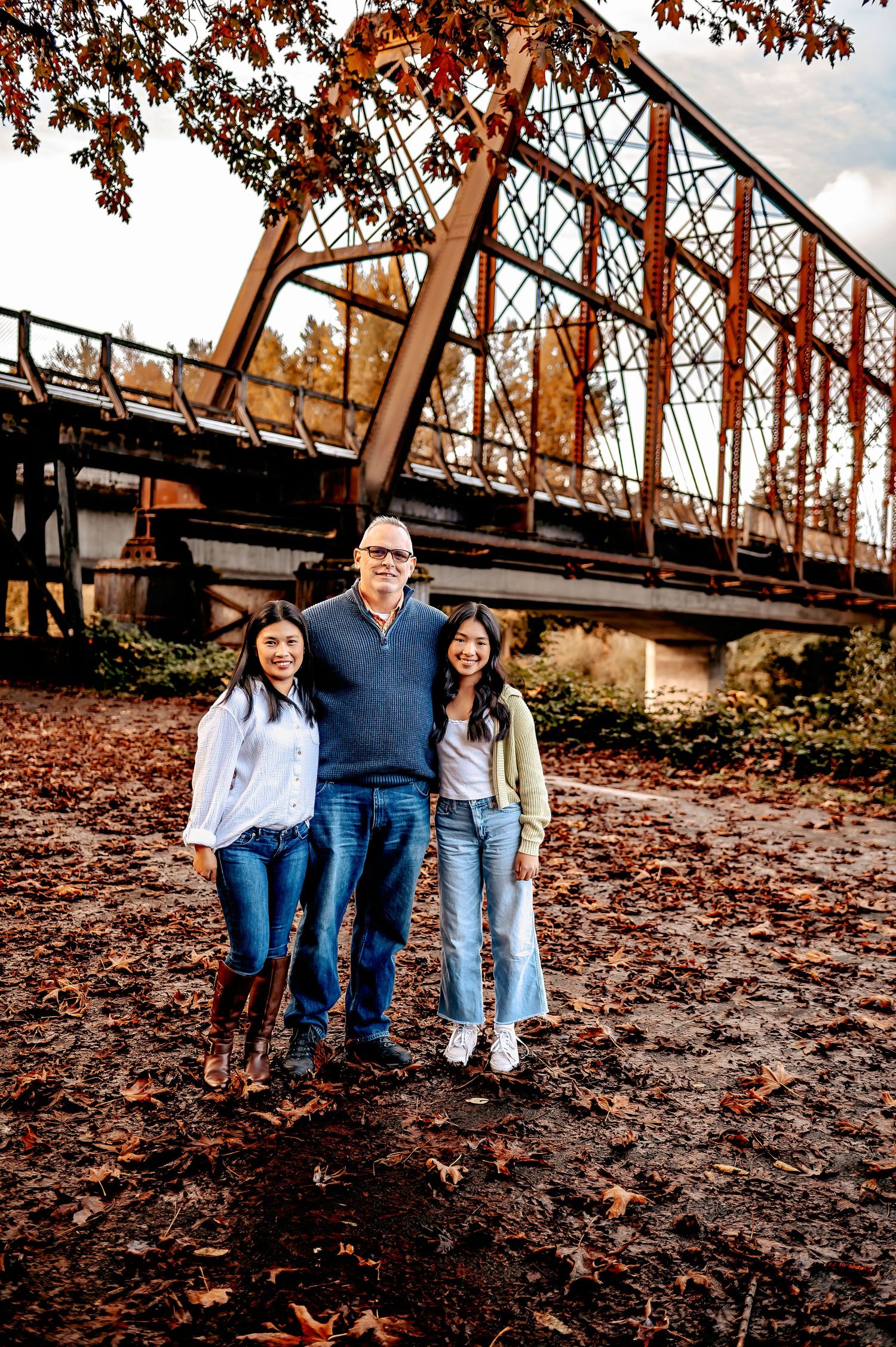 Family sits together on fallen autumn leaves near rustic bridge structure.