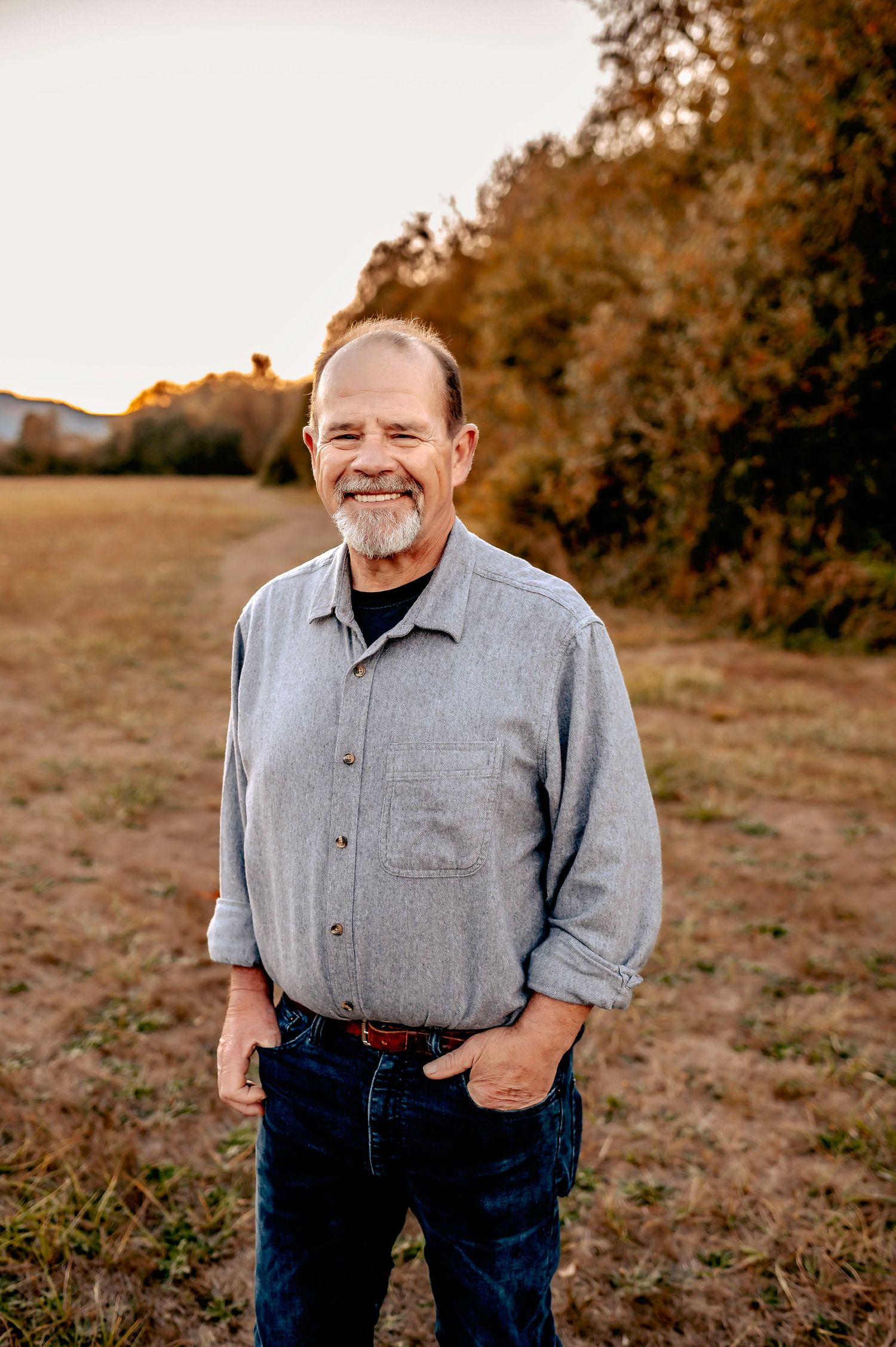 Portrait of person in grey button-up shirt and jeans standing in autumn field at sunset.