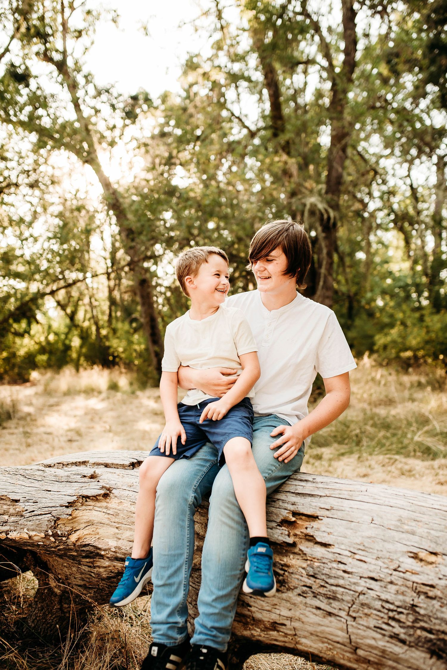 A parent and child share a tender moment while sitting together on a fallen log in a sunlit forest setting.