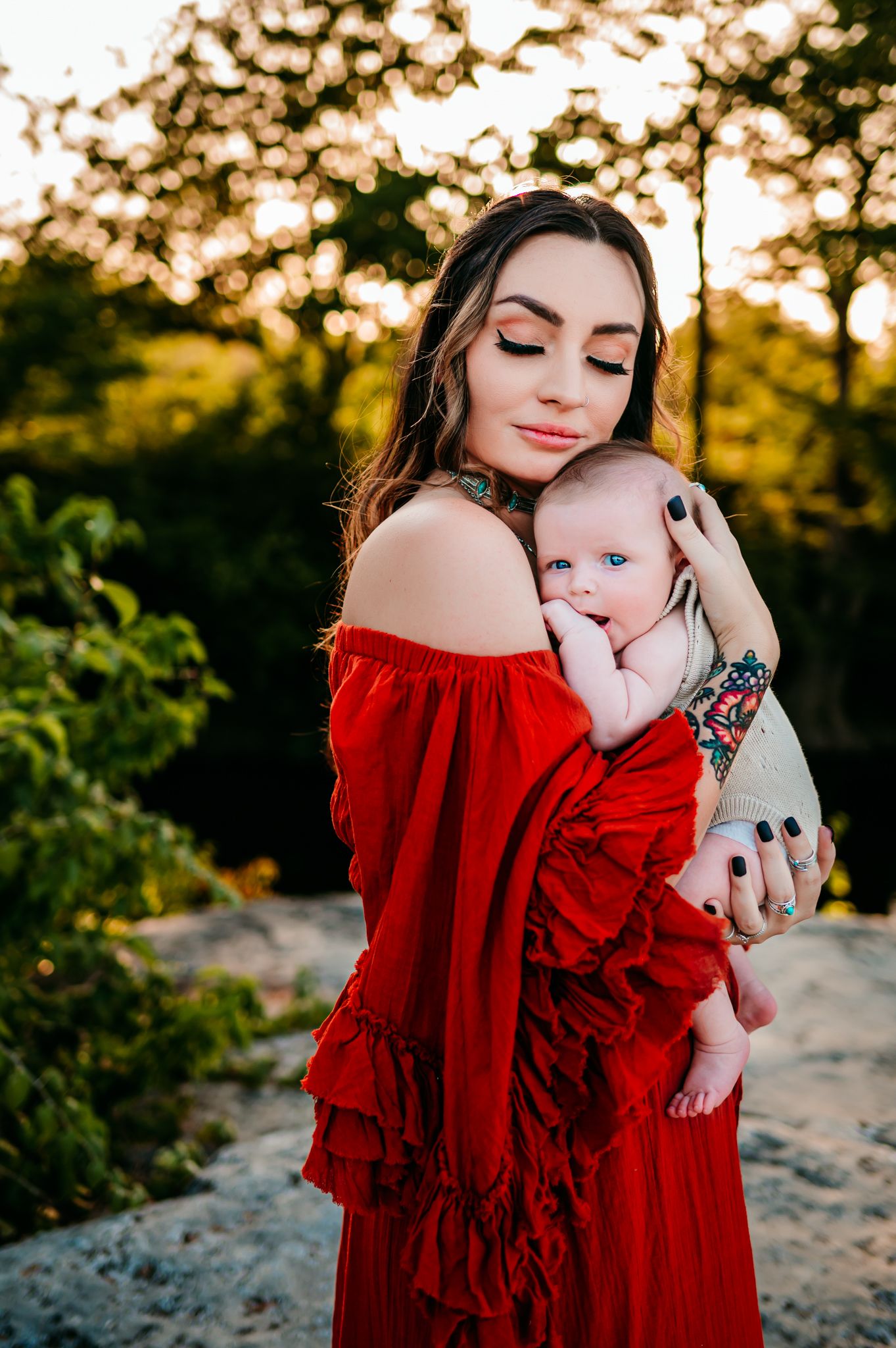 Mother in flowing red dress shares tender moment with baby during outdoor sunset photoshoot.