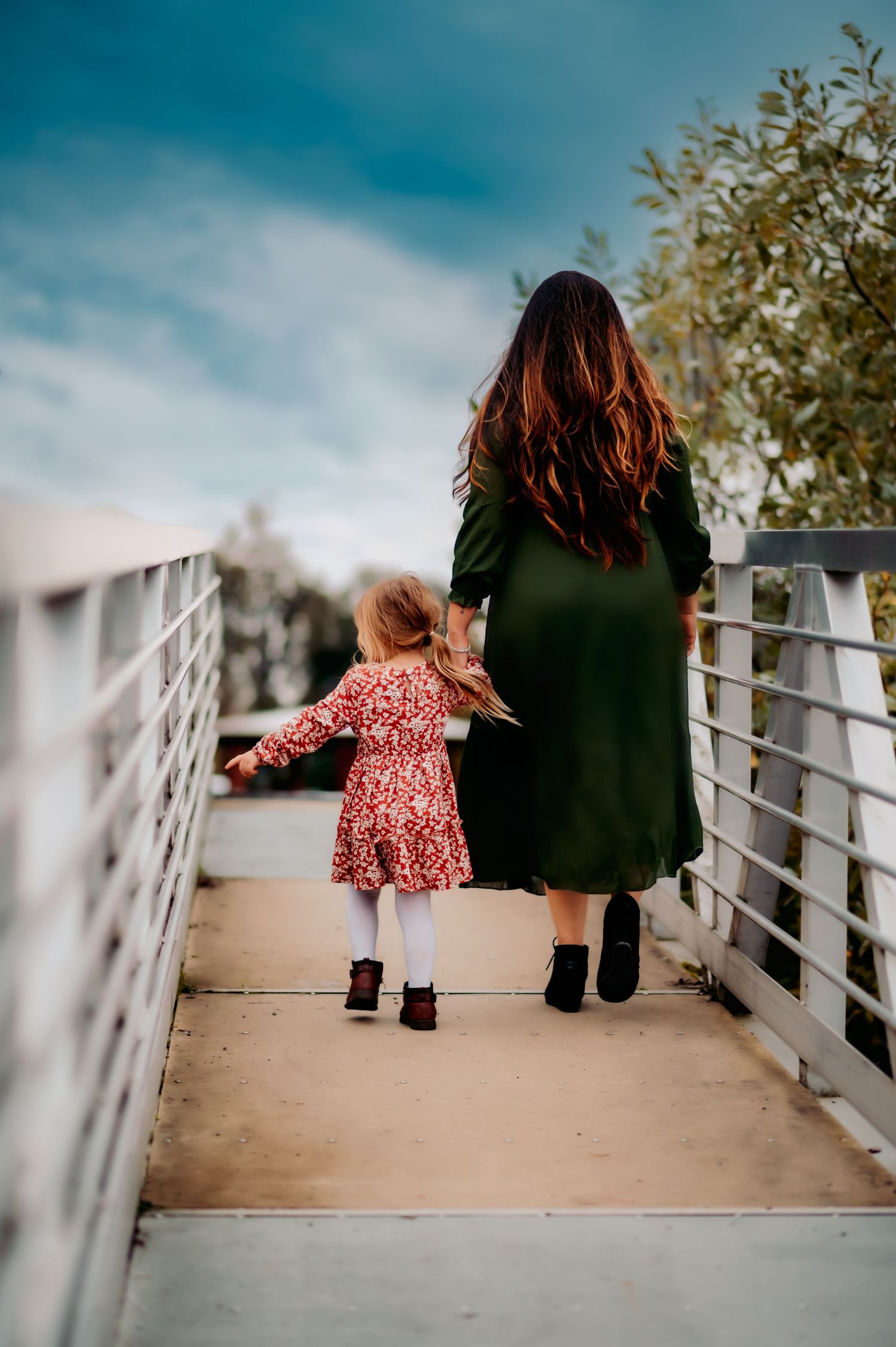 Mother and child walking on concrete path with blue sky and trees.