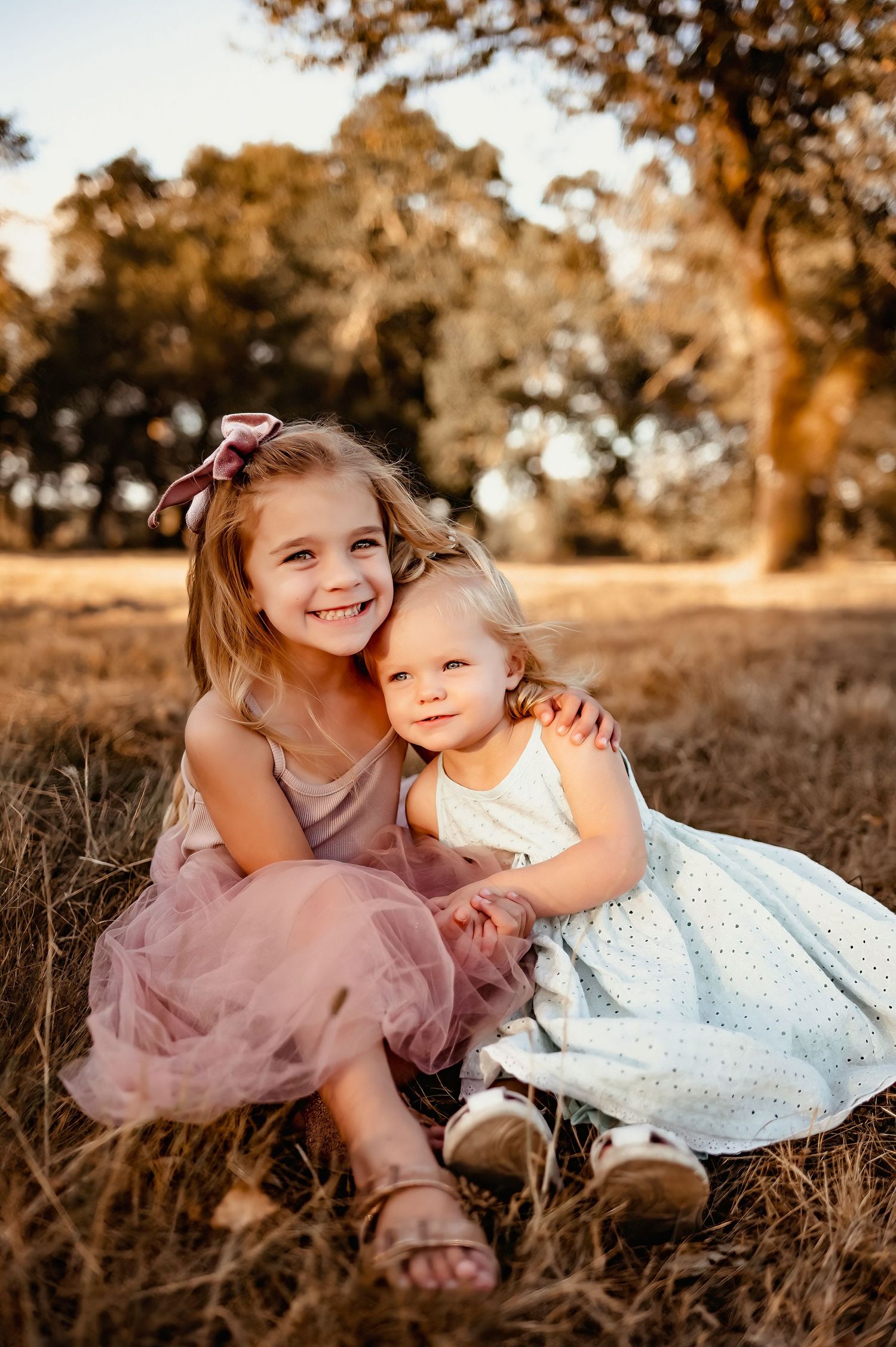 Two children in pink and white dresses share a sweet moment together in a golden sunlit field.