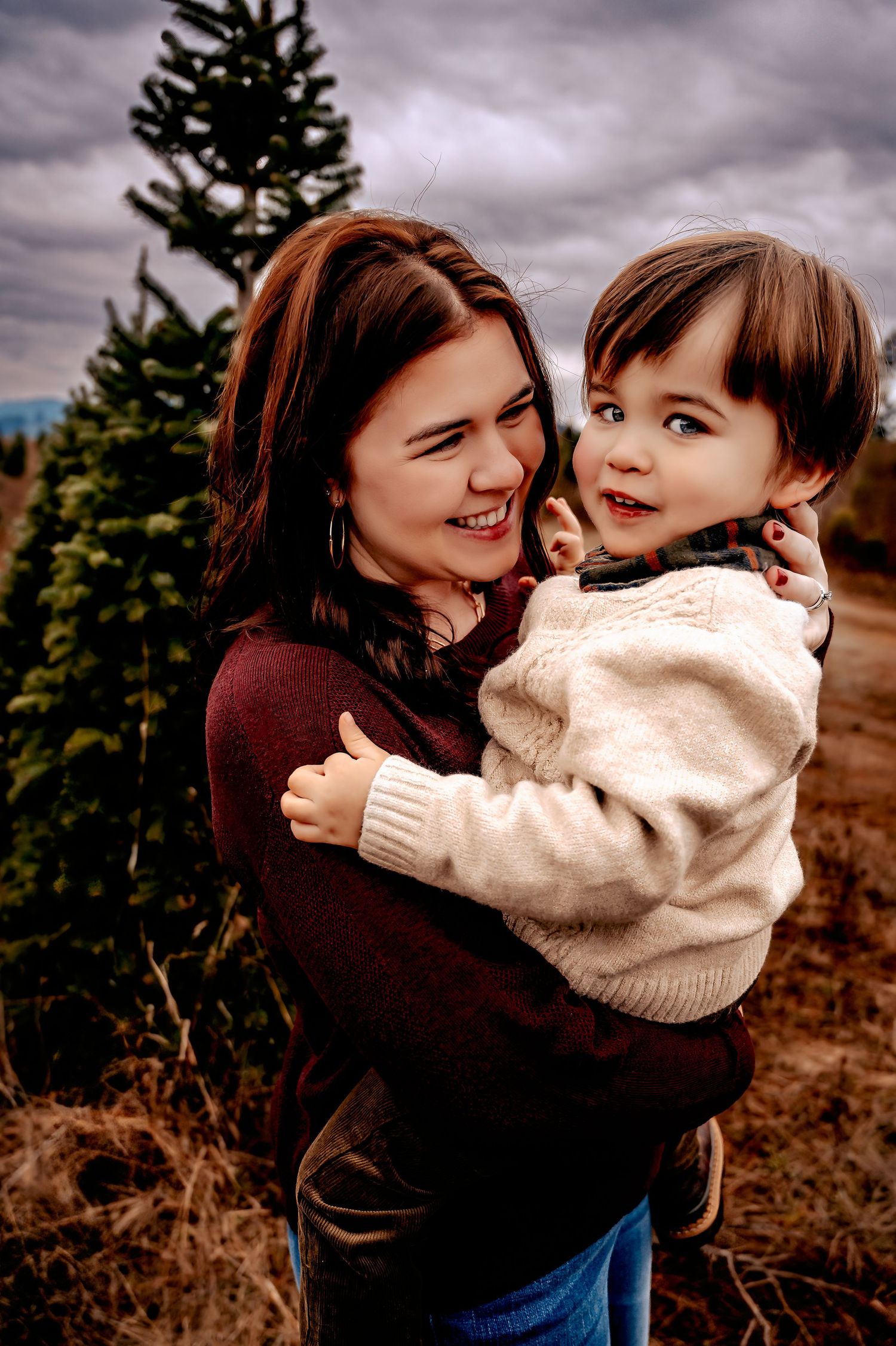 Mother and child share joyful embraces against an evergreen forest backdrop on a cloudy day.