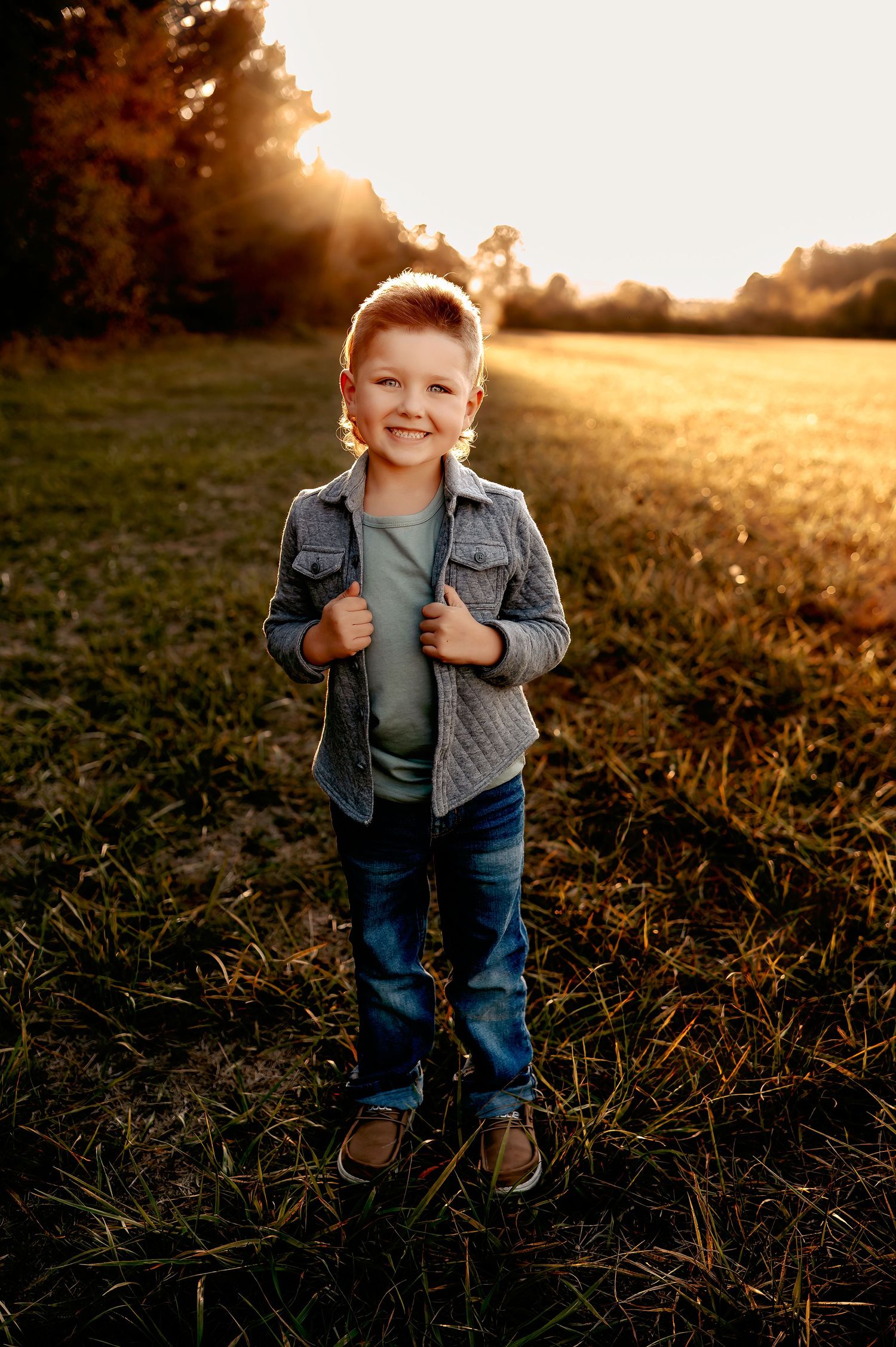 Child in denim jacket and jeans smiling while standing in sunlit field at golden hour.
