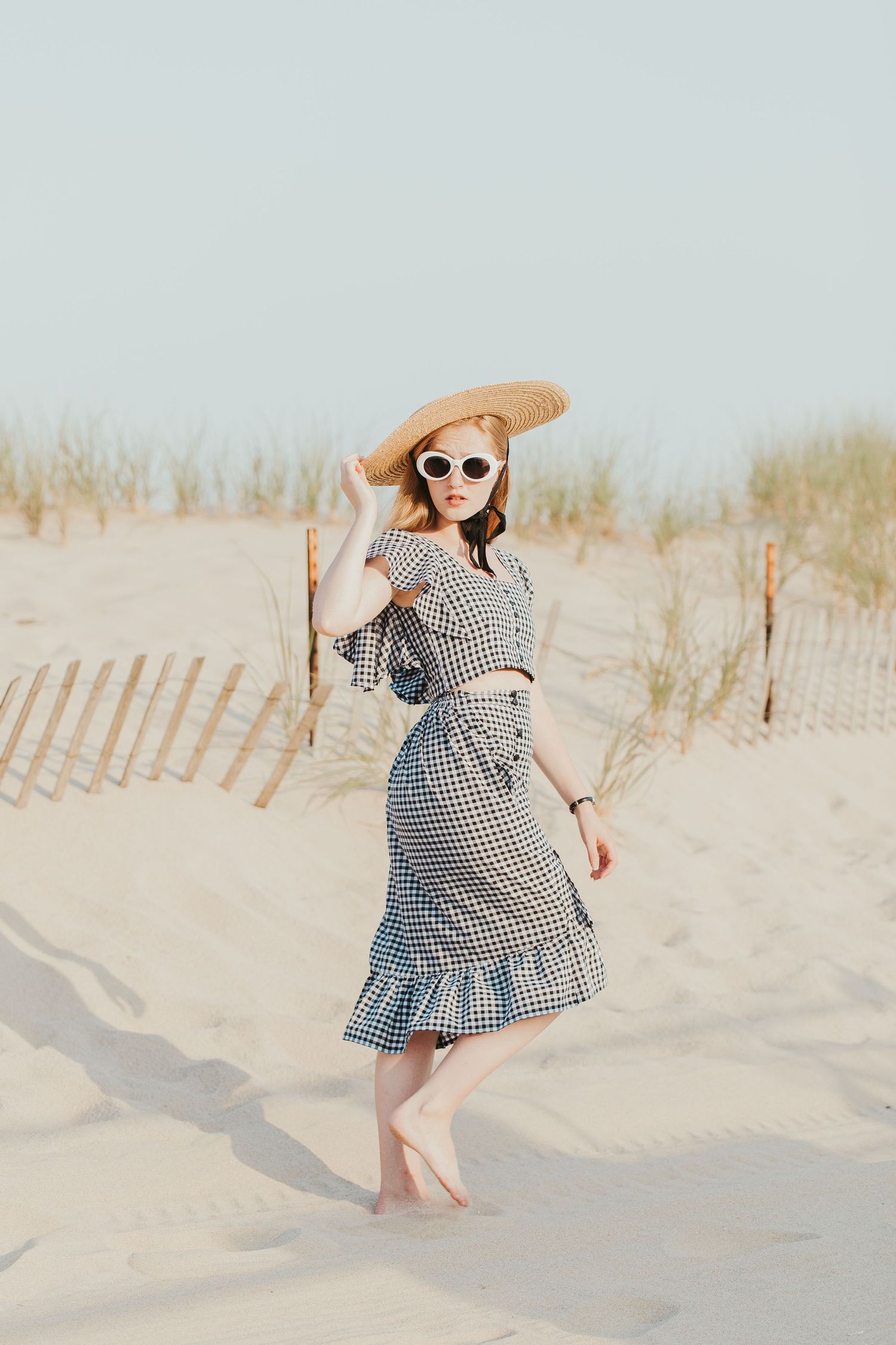 Woman in gingham dress and sunhat posing elegantly on sandy beach dunes during golden hour.