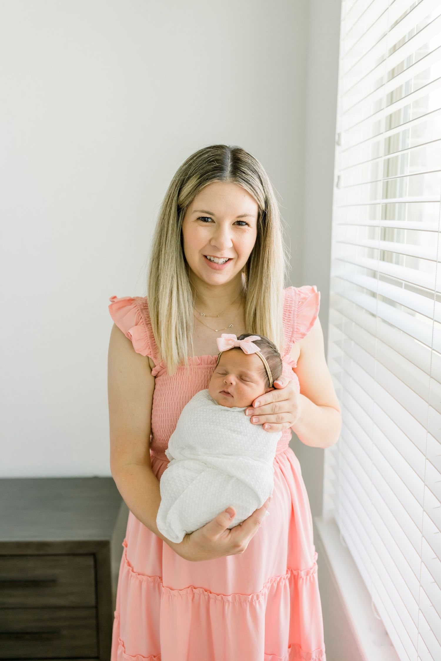 A woman in a pink dress holds her sleeping newborn wrapped in white by a window.