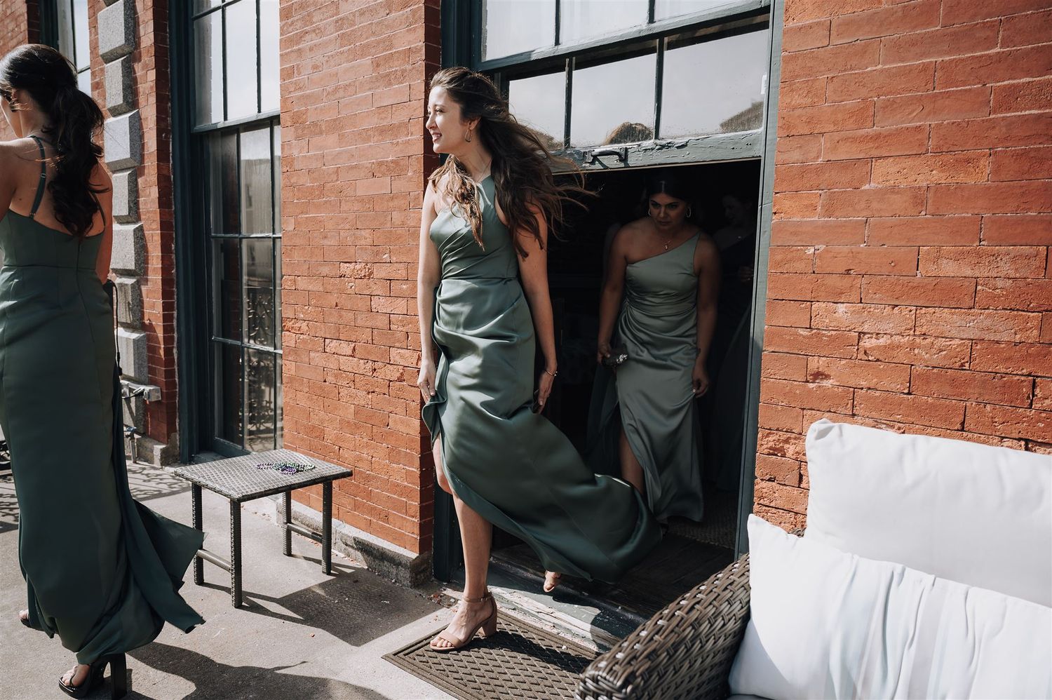 Woman in sage green dress stepping onto sunlit balcony from brick building.