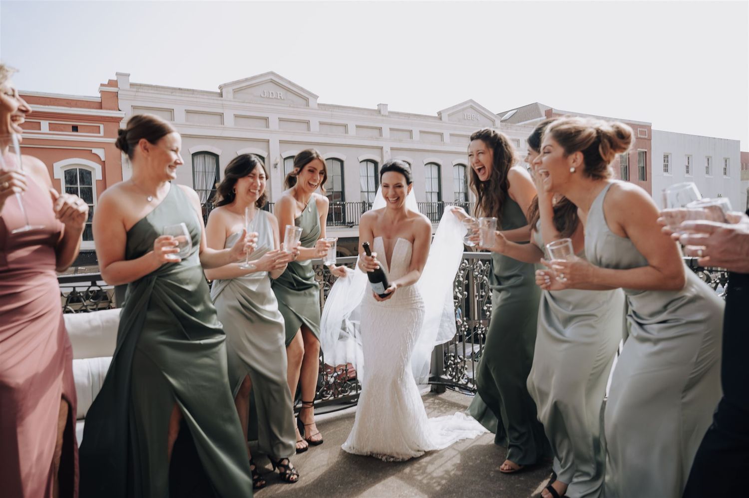 Bridesmaids in sage dresses surround bride in white gown on rooftop terrace.