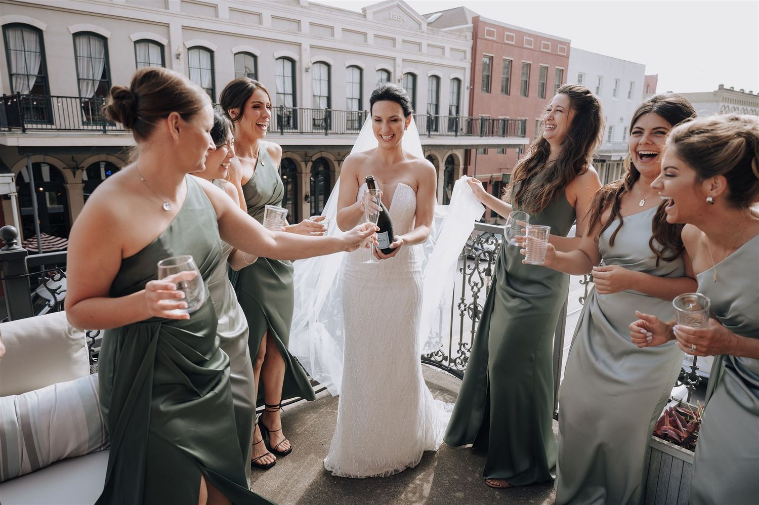 Wedding party in sage green bridesmaid dresses gather on a balcony overlooking historic brick buildings.