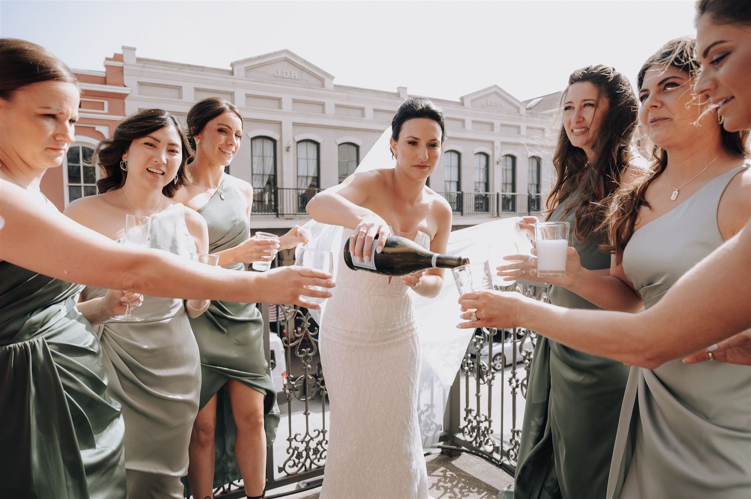 Wedding party pouring champagne on sunny balcony with historic architecture in background.