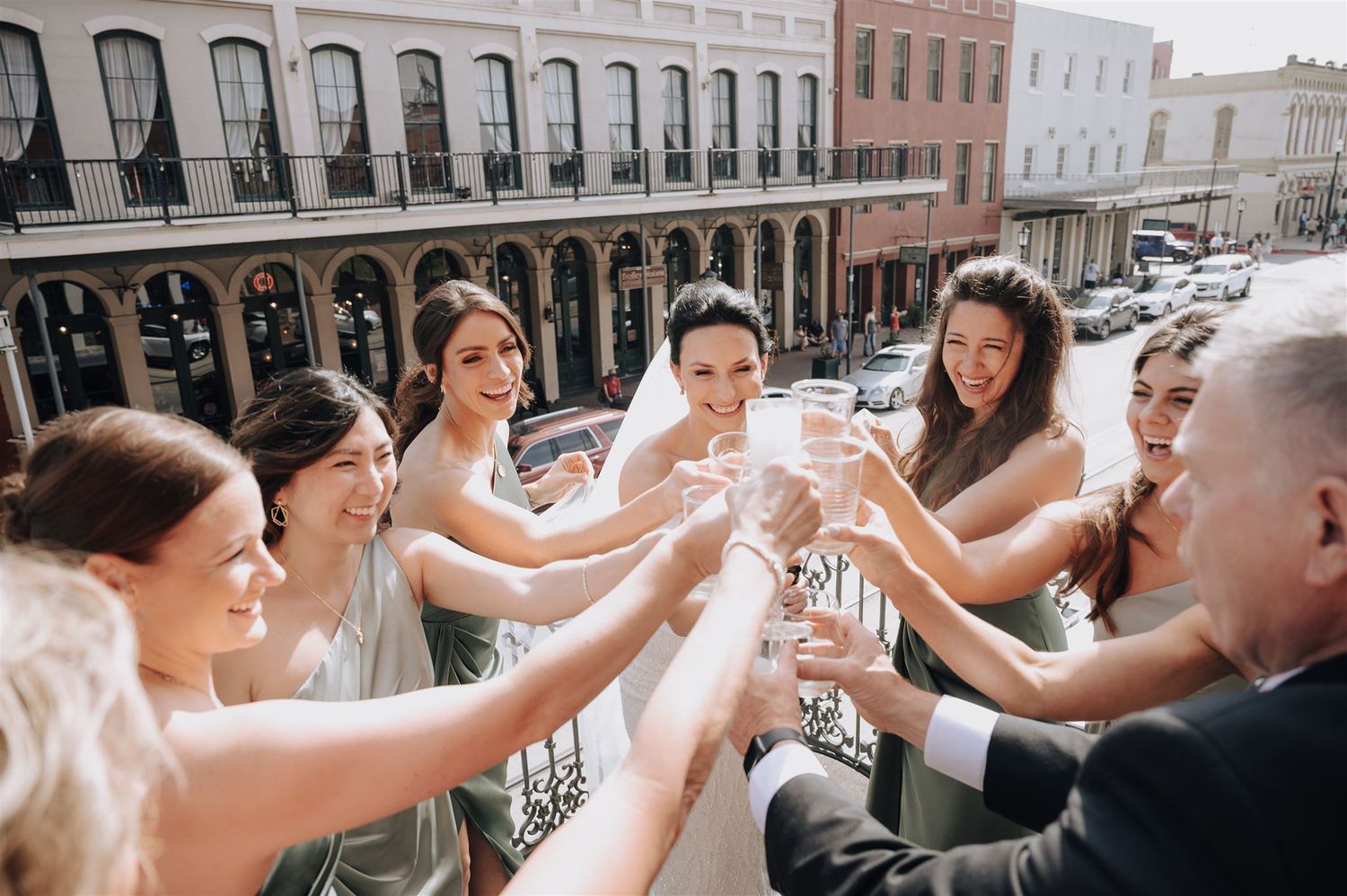 Wedding party toasts with champagne on balcony overlooking historic downtown buildings.