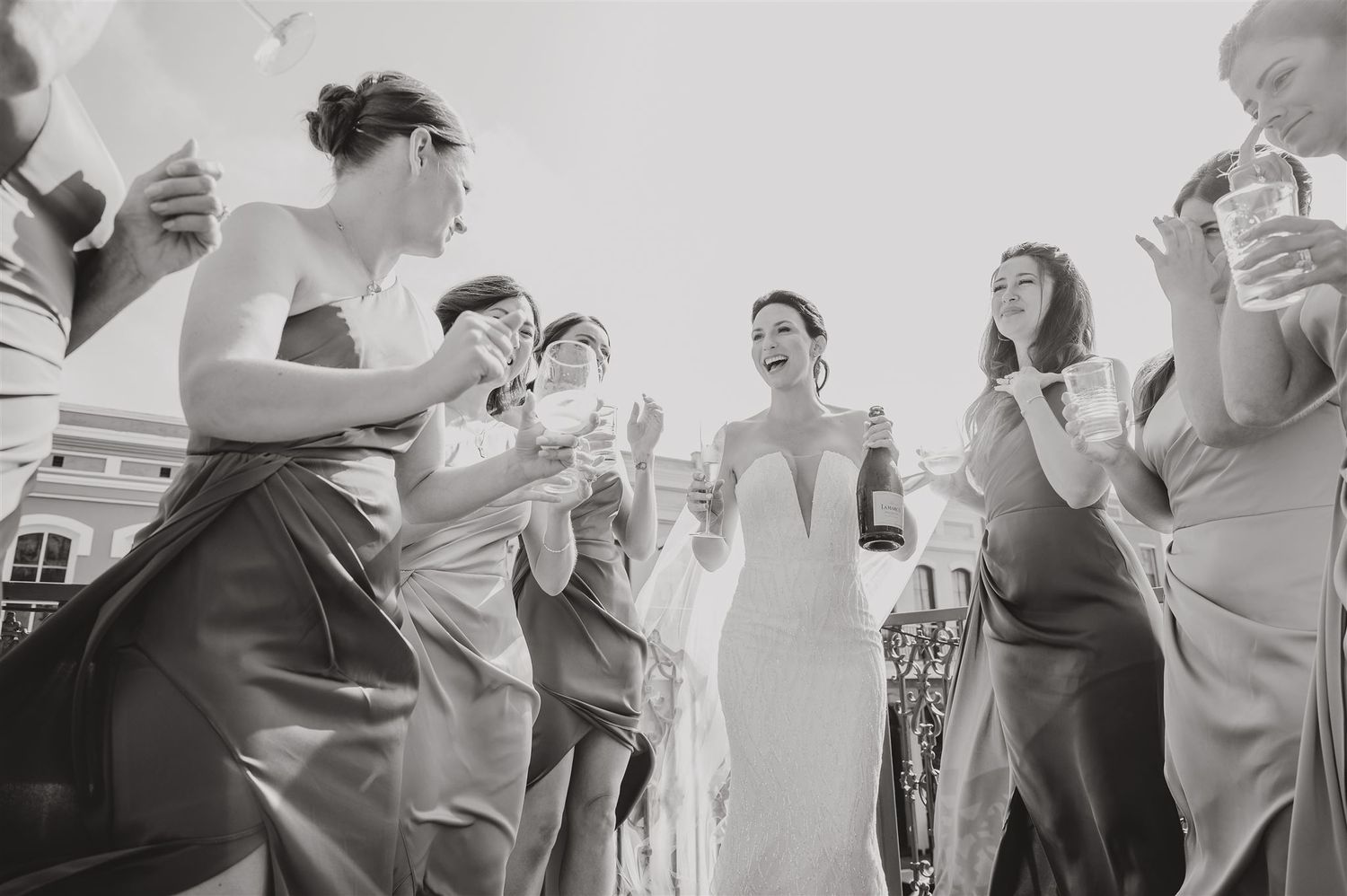 Wedding party celebrates joyfully on sunny balcony with historic architecture backdrop.