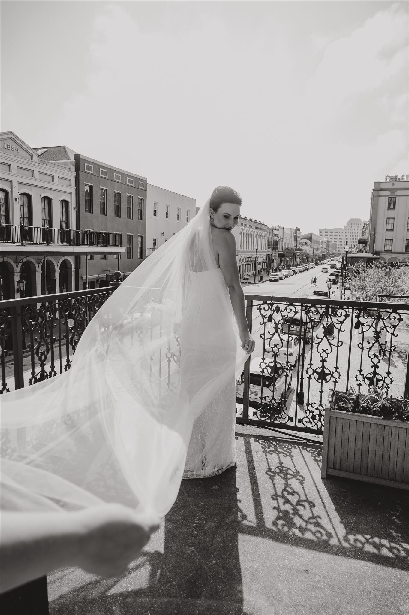 Flowing white wedding veil catches breeze on historic building balcony overlooking city street.