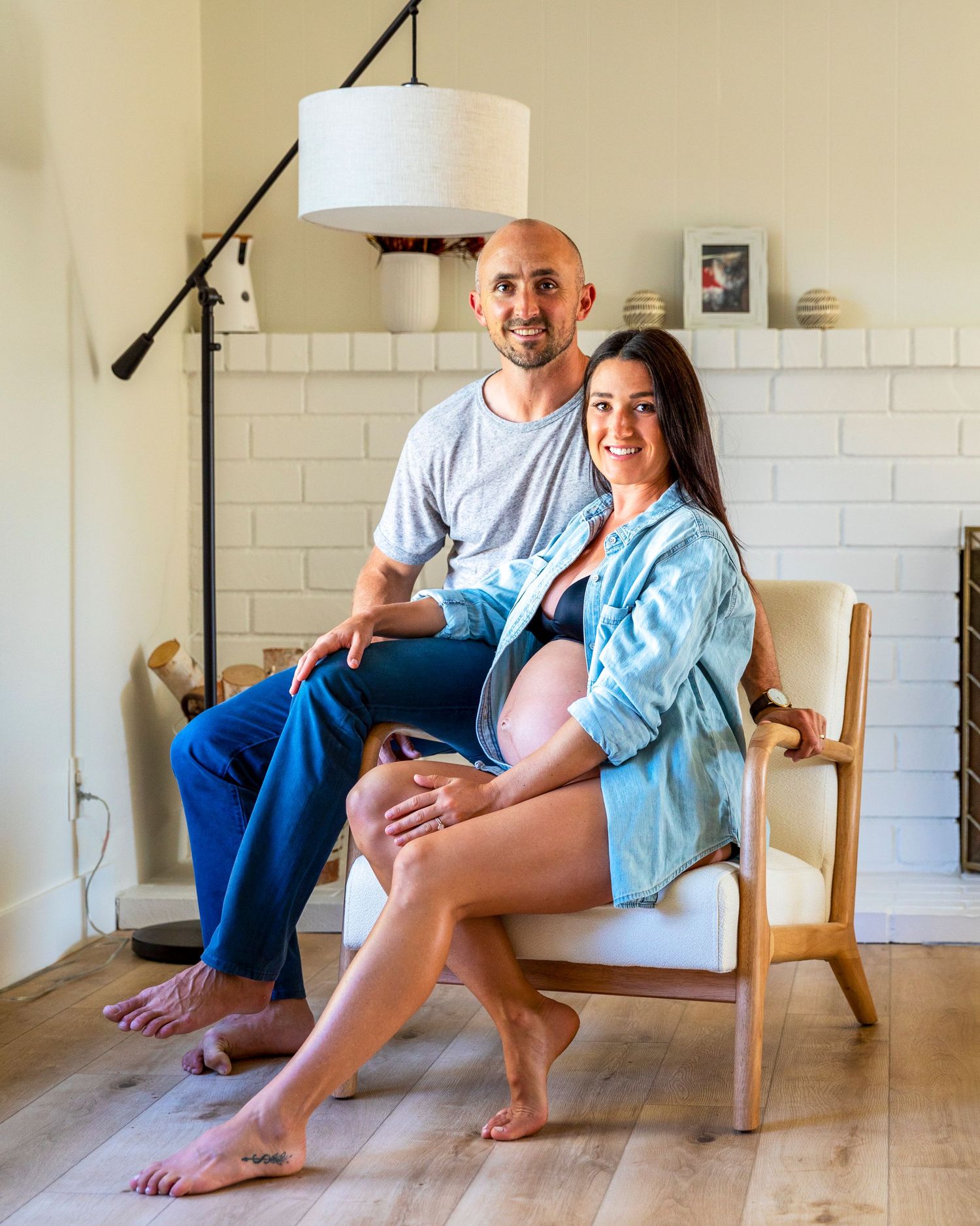 In-home maternity photoshoot wiSmiling couple posing together, pregnant woman seated on chair with partner beside her in bright living room.