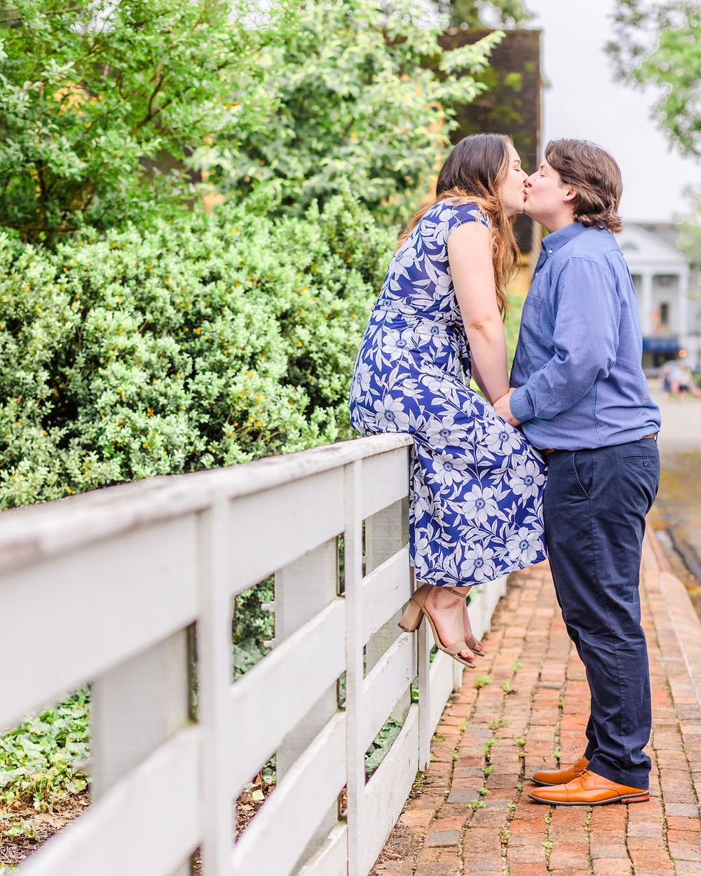 Couple embraces romantically by white fence surrounded by greenery.