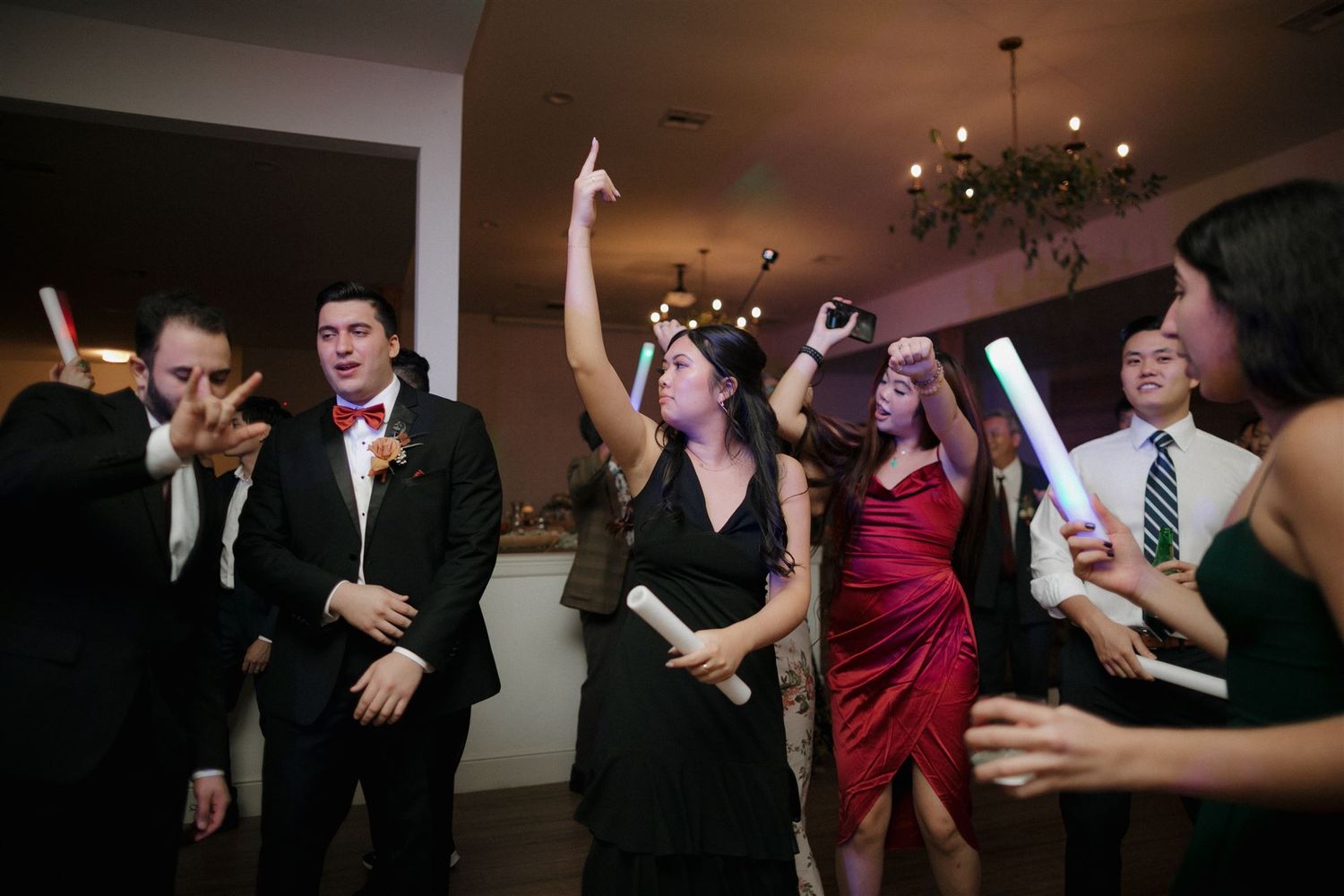 Partygoers dancing enthusiastically at an indoor wedding celebration with chandeliers.
