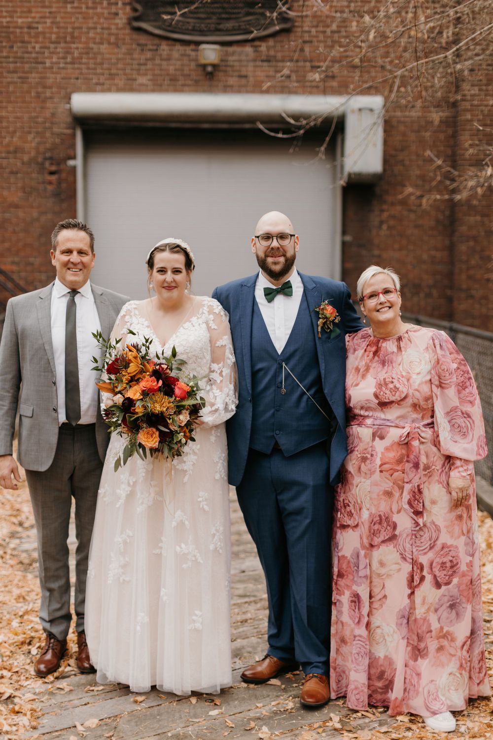 Bride in lace gown and groom in navy suit pose with parents in floral dress and gray suit at fall wedding.