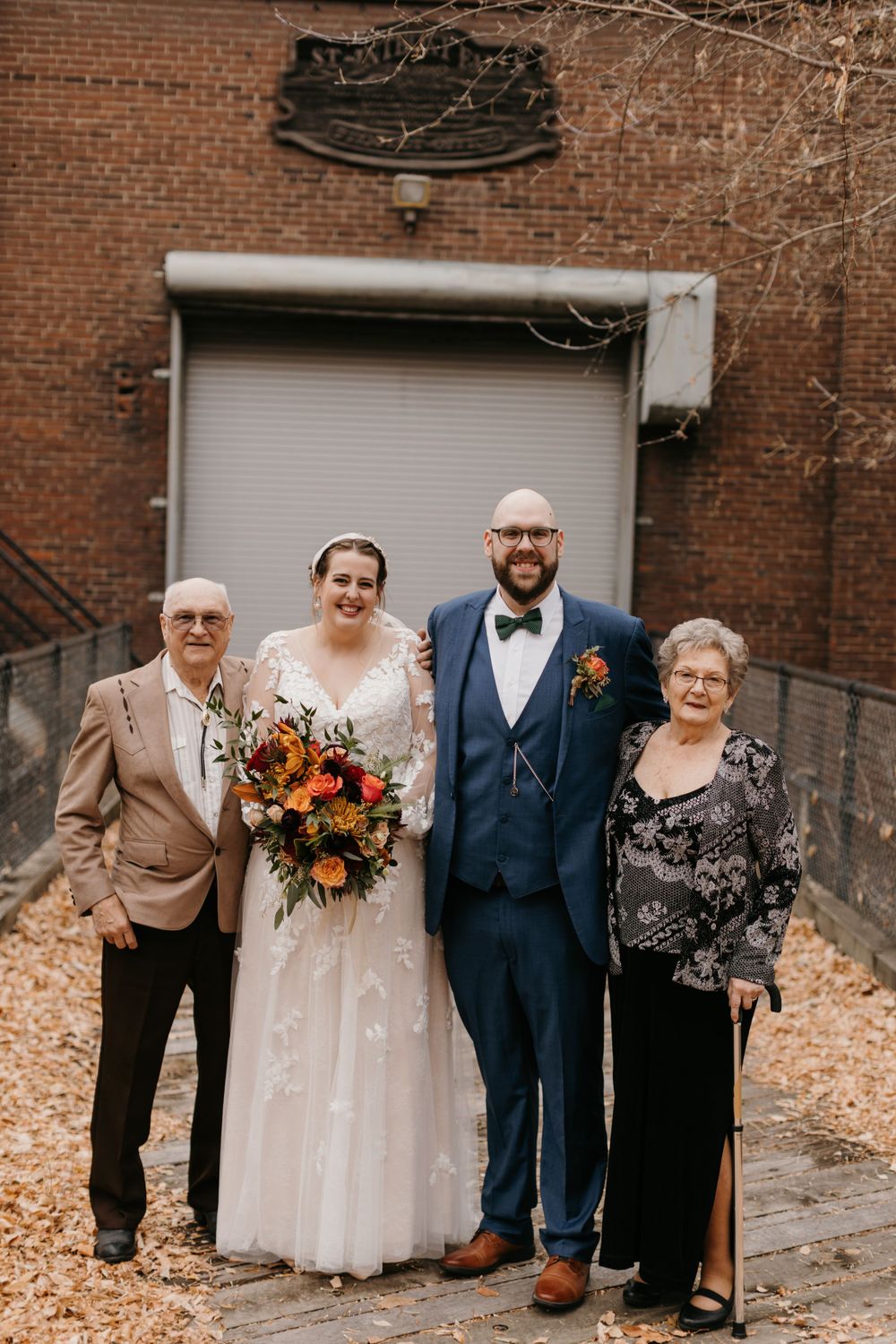 Bride and groom pose with elderly grandparents outside brick building surrounded by fallen autumn leaves.