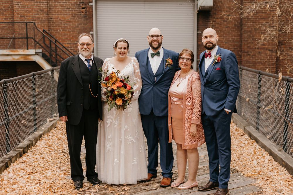 Wedding party of five posing outdoors in fall foliage, bride holding autumn bouquet with groom in navy suit.