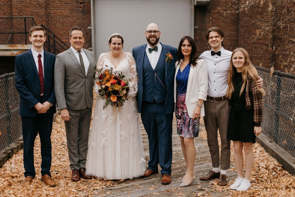 Wedding party of seven smiling outdoors among autumn leaves beside rustic brick building backdrop.