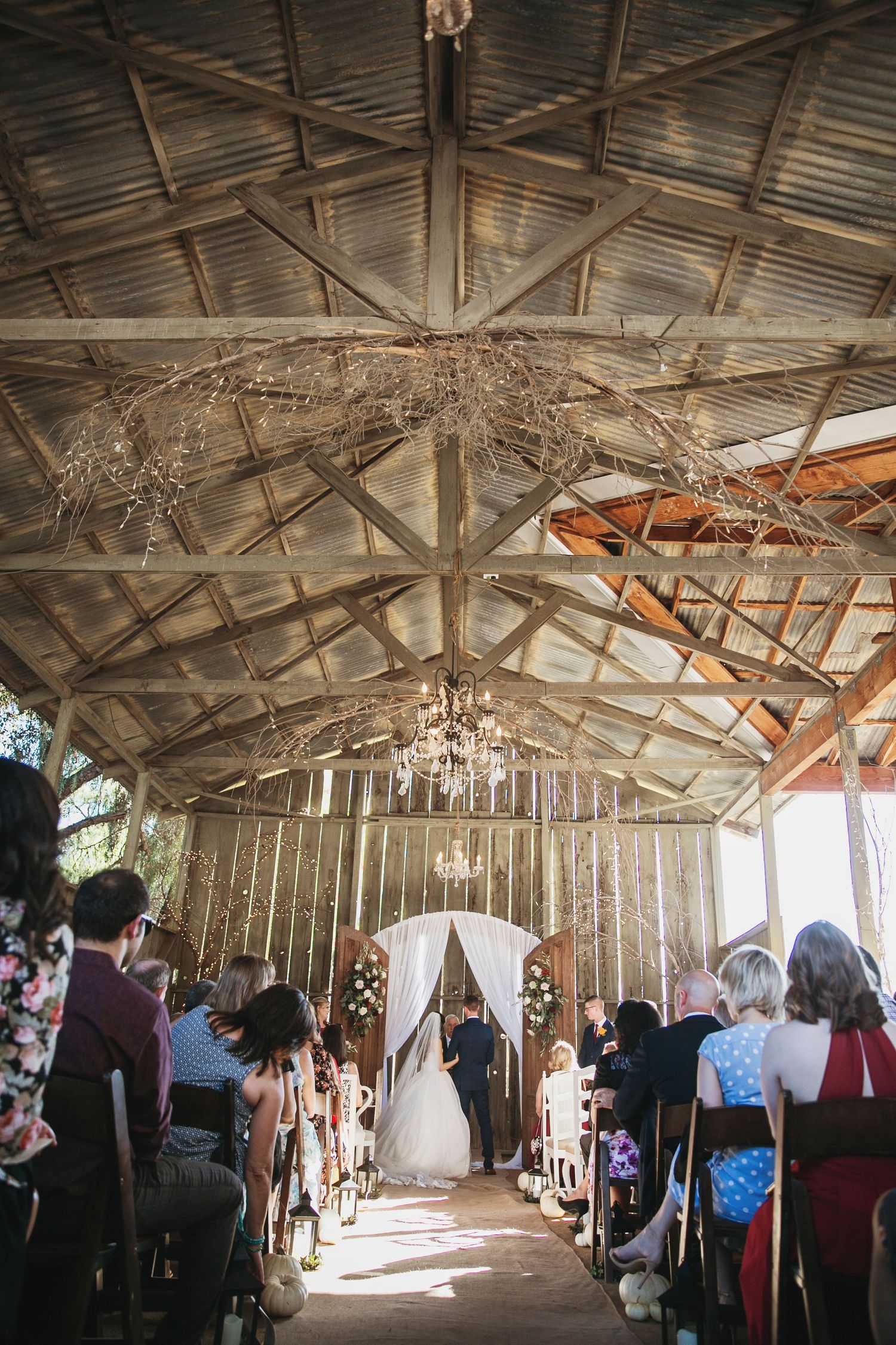 A rustic barn wedding ceremony with crystal chandeliers, wooden beams, and guests seated in rows beneath a vaulted ceiling.