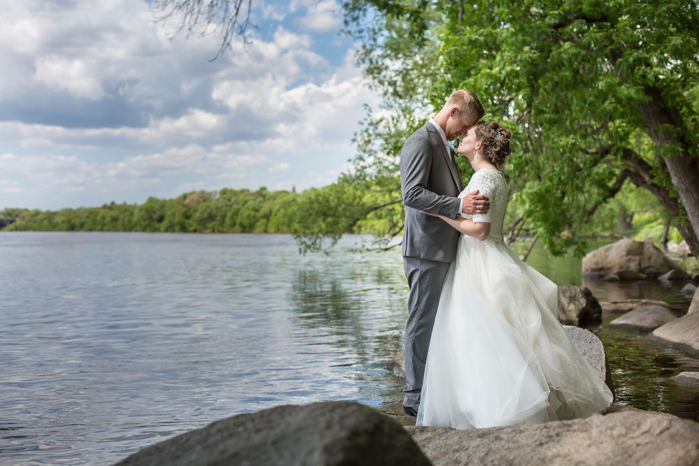 Bride and groom share a romantic moment by a serene lakeside, surrounded by lush green trees on a sunny day.