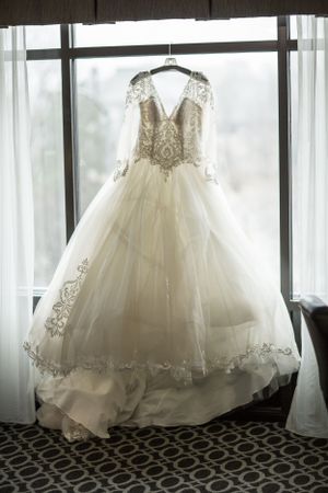 Elegant white ball gown wedding dress with silver lace embroidery and long sleeves hanging by a window.