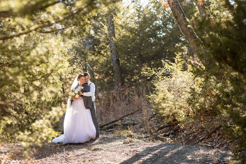 Bride and groom share a romantic embrace surrounded by lush green trees in a sunlit forest setting.