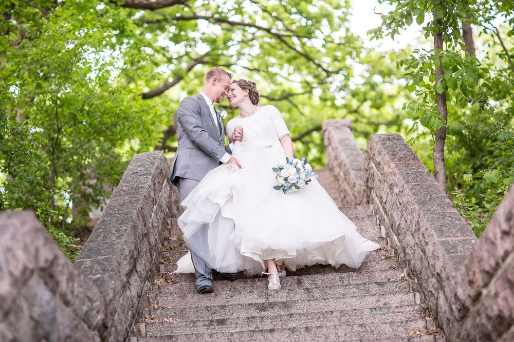 Bride and groom share a romantic moment on stone bridge steps surrounded by lush green trees on their wedding day.
