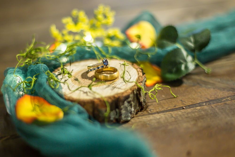 Gold wedding rings displayed on a rustic wood slice with teal ribbon and yellow floral accents.