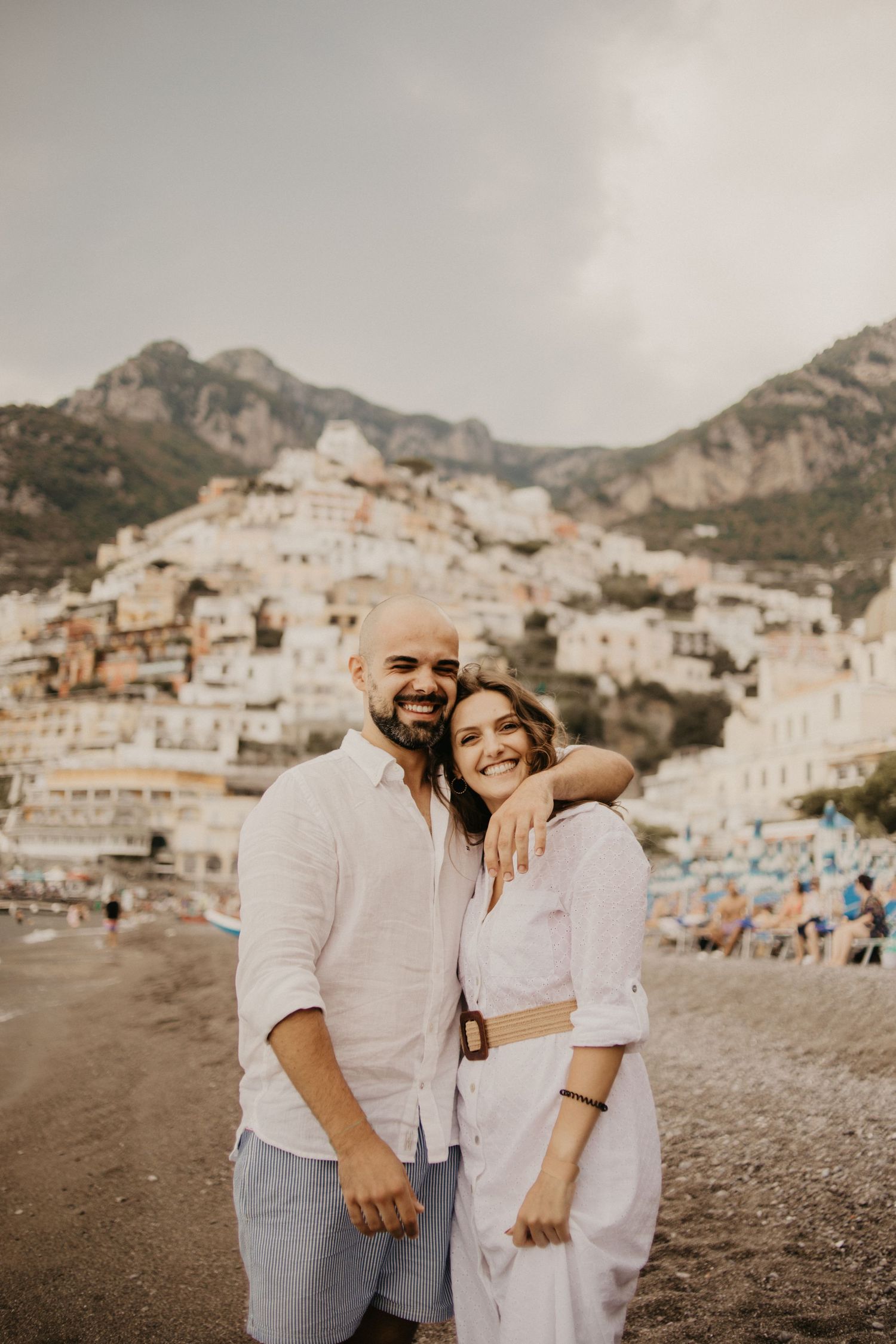Couple in white outfits embracing on beach with iconic Positano coastal town and mountains in background.