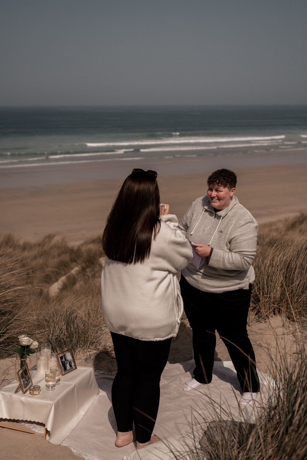 Two people exchange vows on a sandy beach dune with ocean waves in the background on a clear day.
