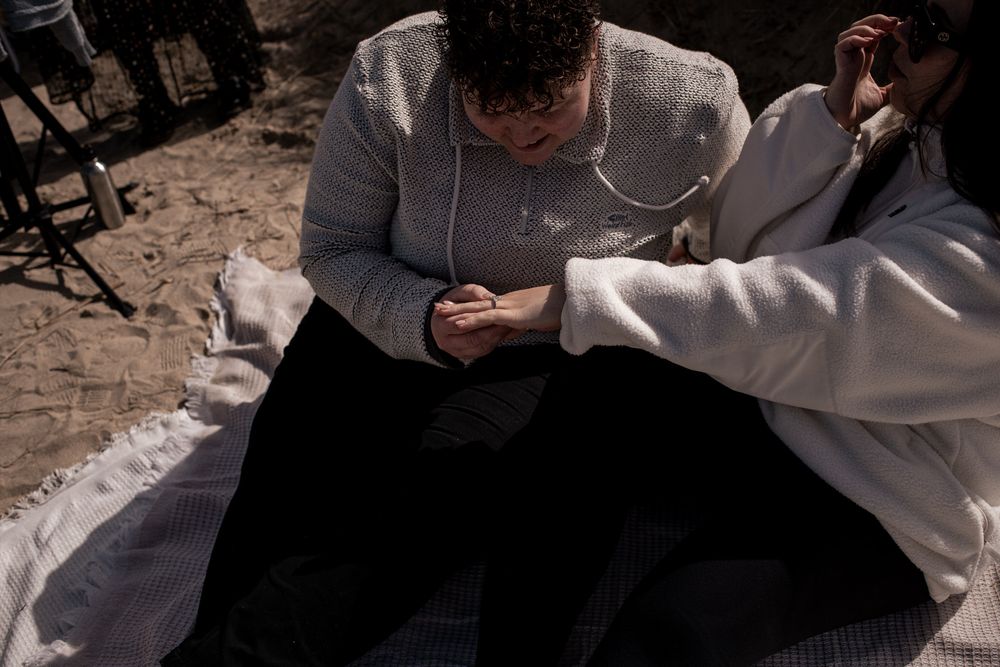 Two people sitting on a blanket on sandy ground, one examining an object held in their hands outdoors.
