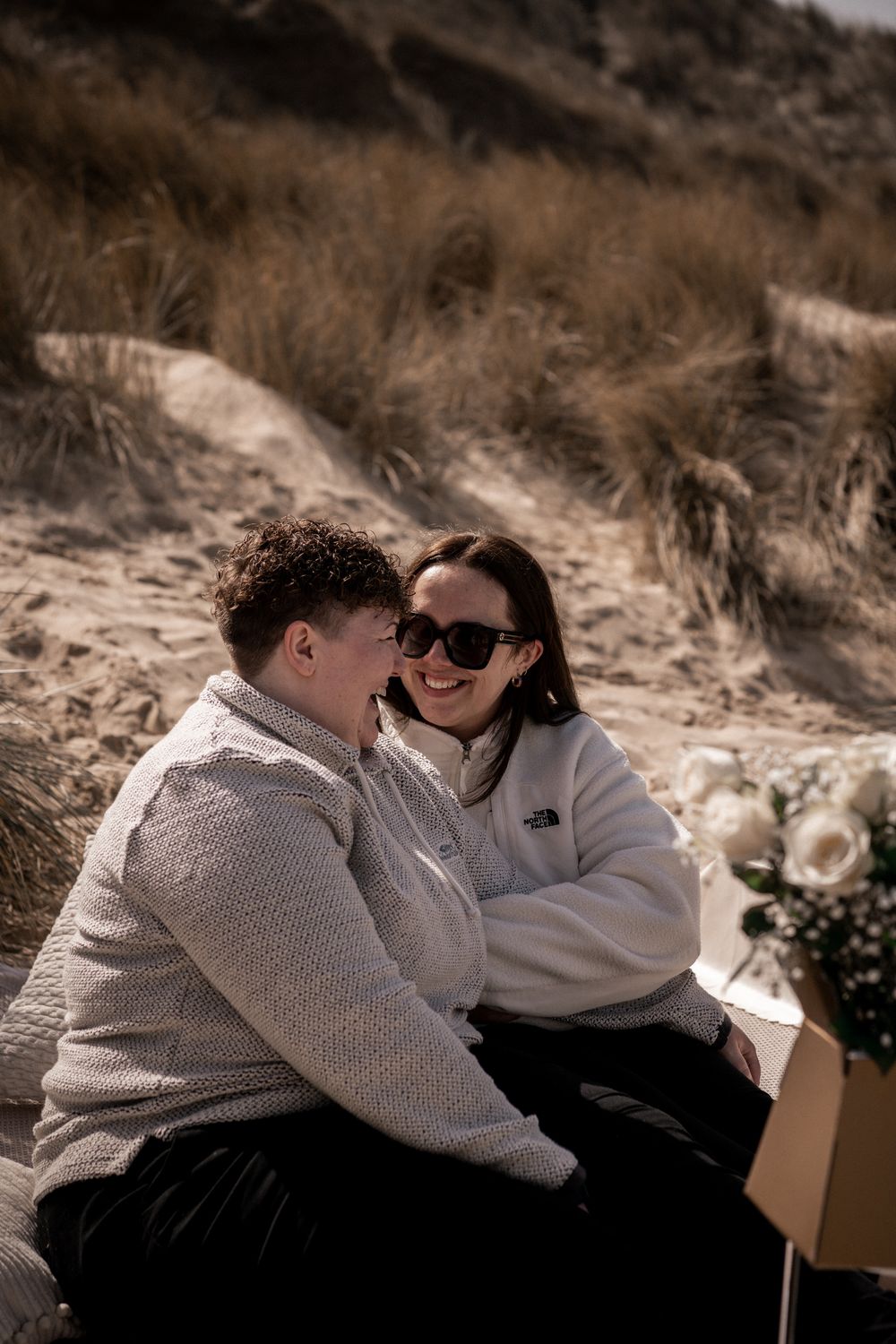 Couple laughing together on sandy beach dunes, wearing cozy white sweaters with flowers nearby.